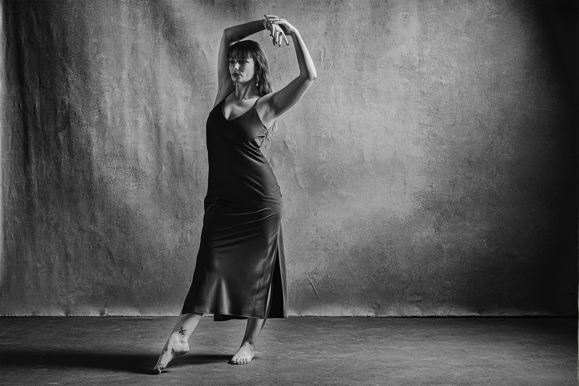 A woman in a long black satin dress is posing with her arms raised above her head in front of a textured gray wall.