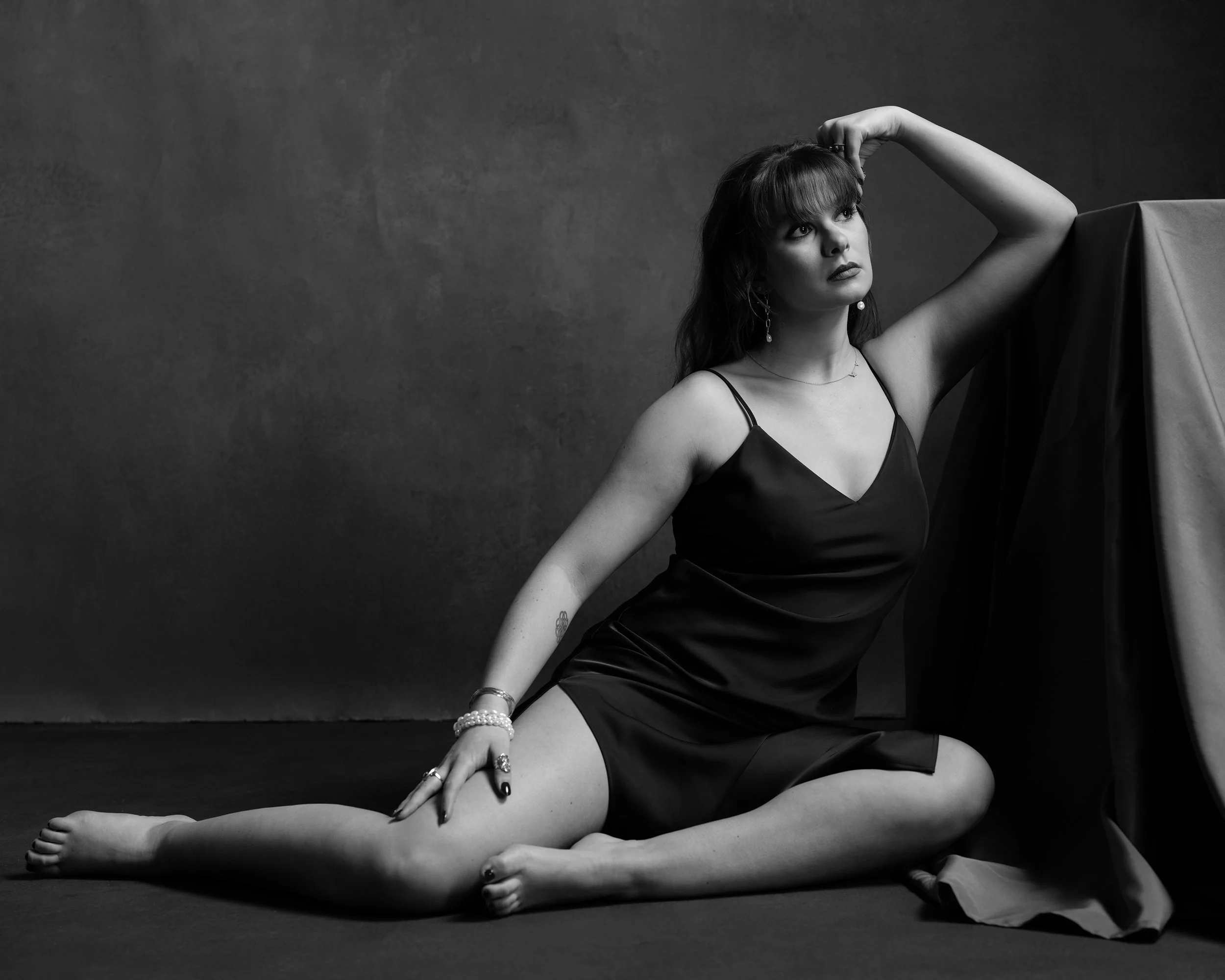 A black and white photo of a woman in a satin slip dress, sitting on the floor with her legs extended and one arm resting on a fabric-covered structure, gazing upward with her hand in her hair.