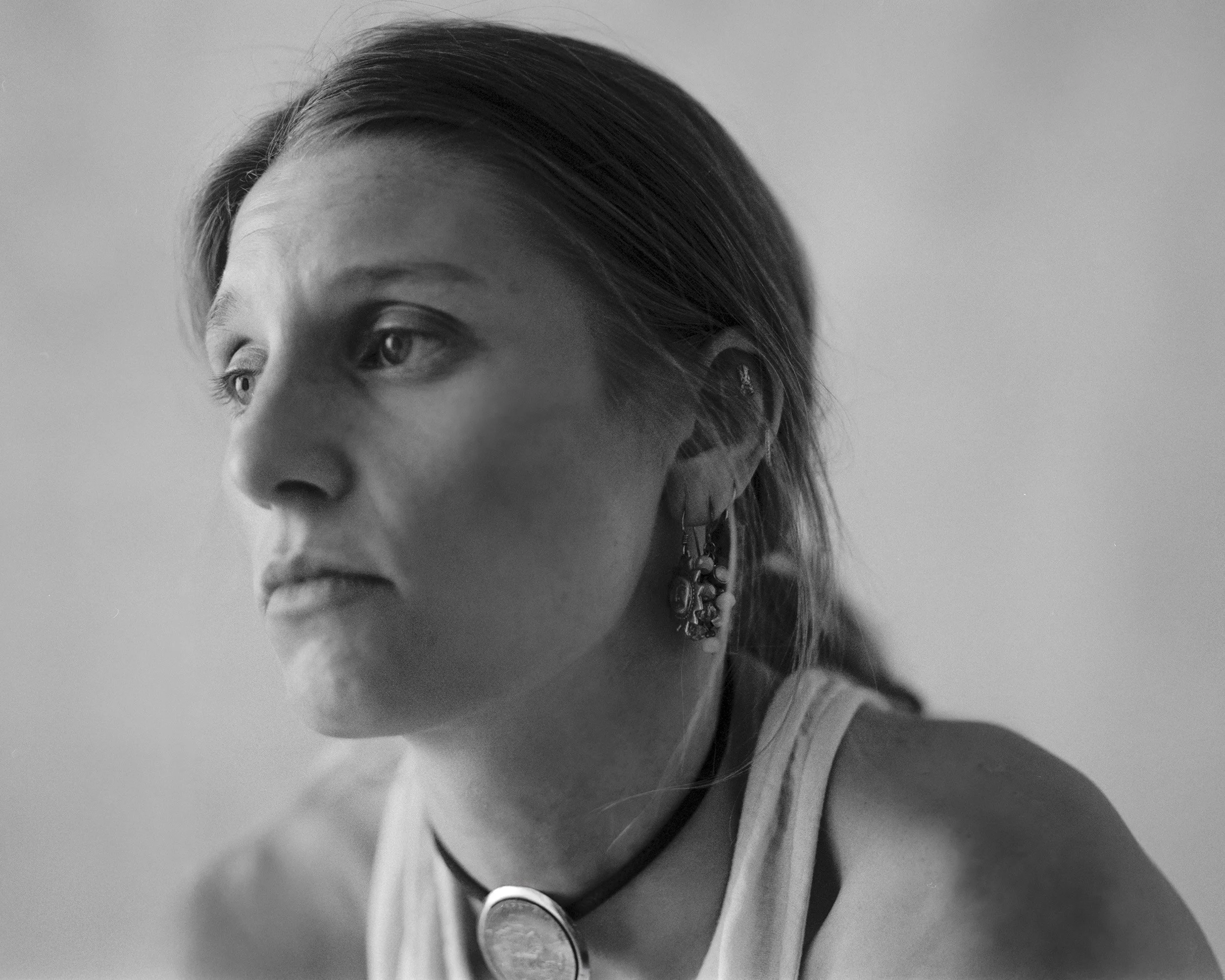 A black and white close-up photograph of a woman with short hair, wearing earrings, a choker, and a top, with her face turned slightly to the left.