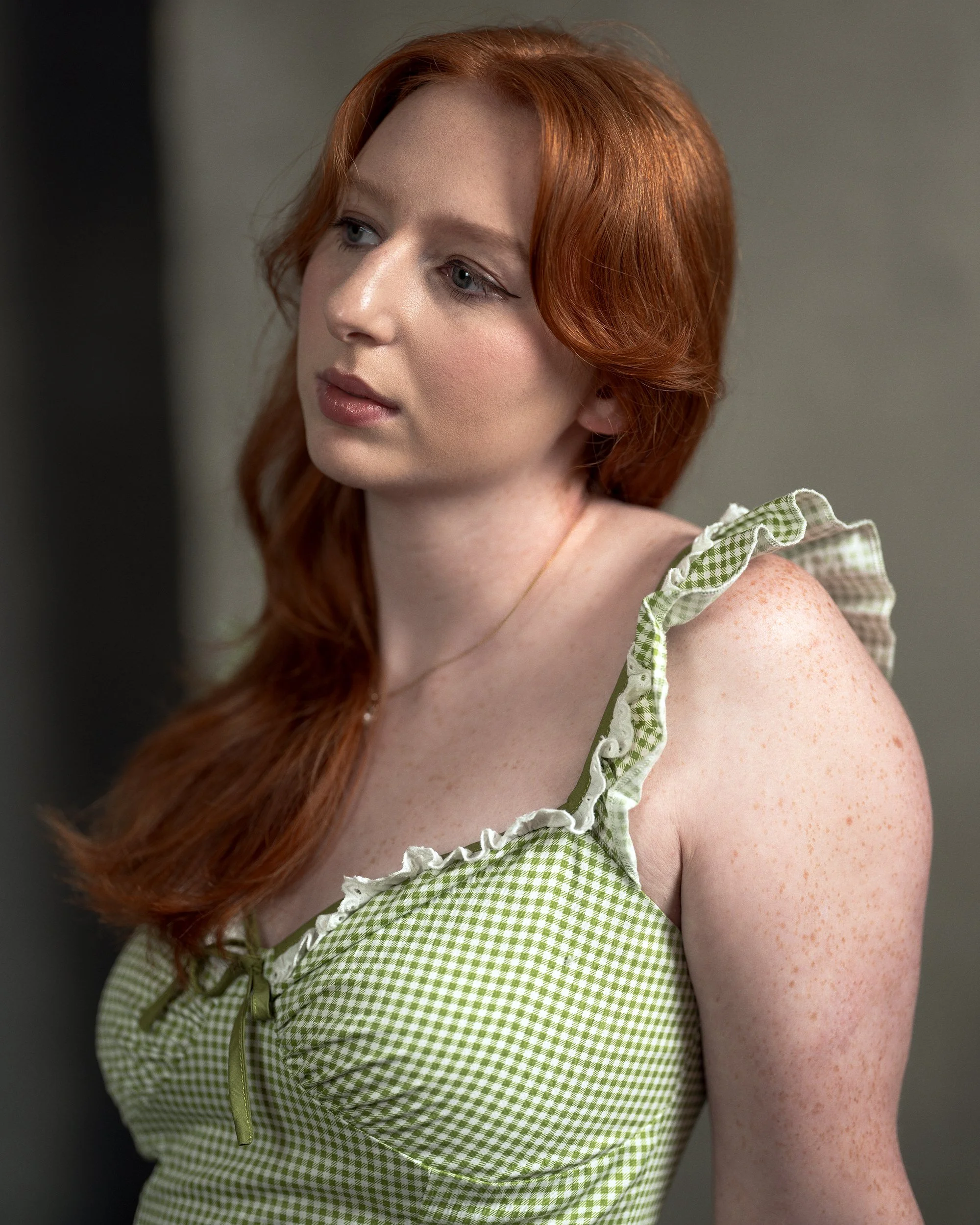 Close-up of a young woman with red hair, freckles, and light makeup, wearing a green and white checkered dress with ruffled straps.