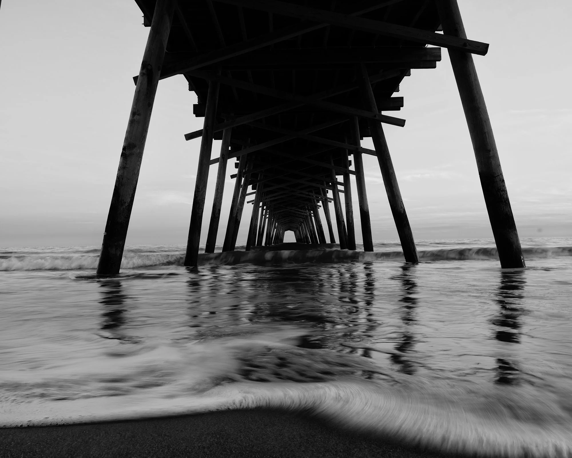Underneath a wooden pier extending into the ocean, with a view from beneath, waves lapping the shore and a cloudy sky above.