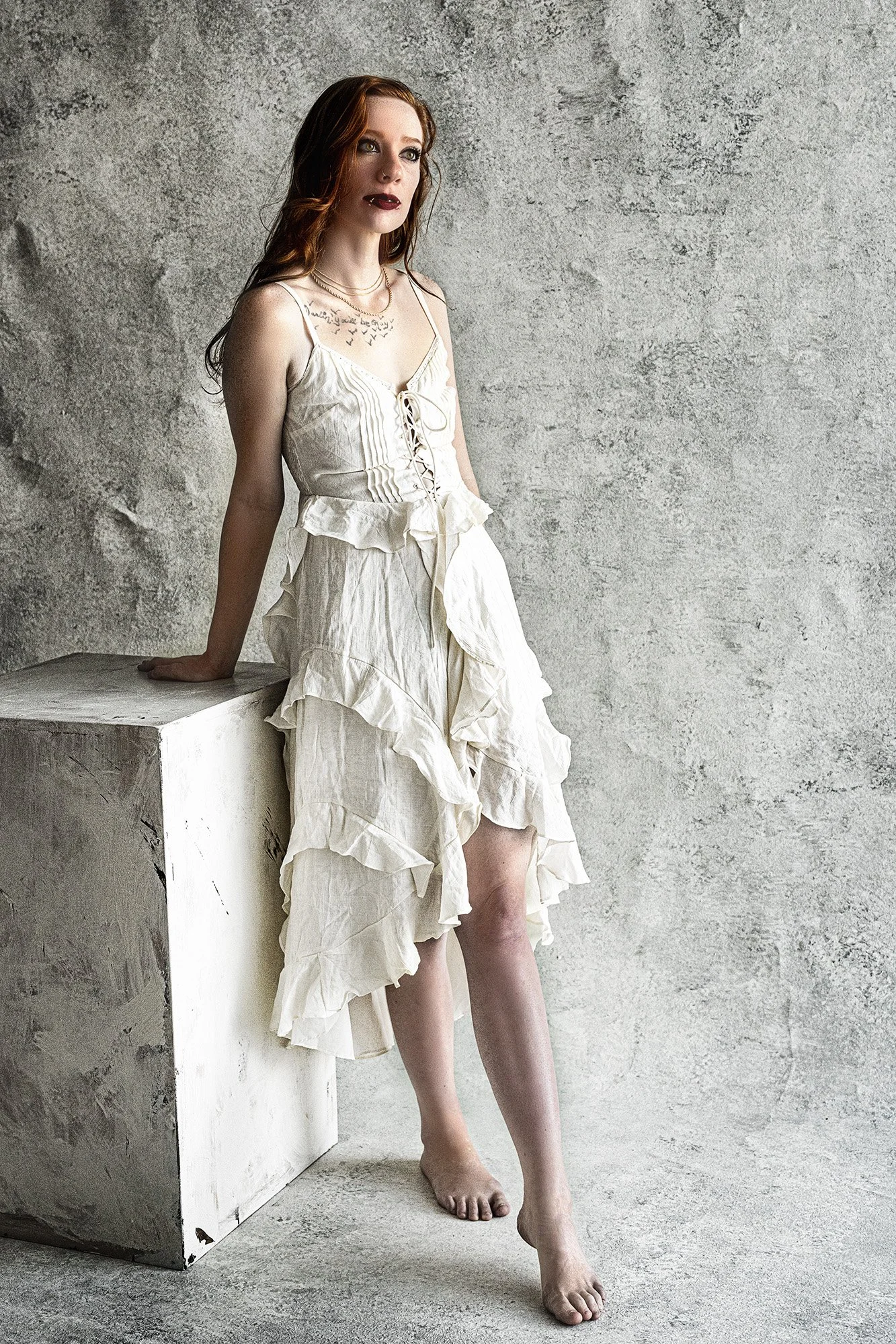 A woman with red hair and dark lipstick standing barefoot next to a white block against a textured gray wall, wearing a cream-colored ruffled dress with a lace-up bodice.