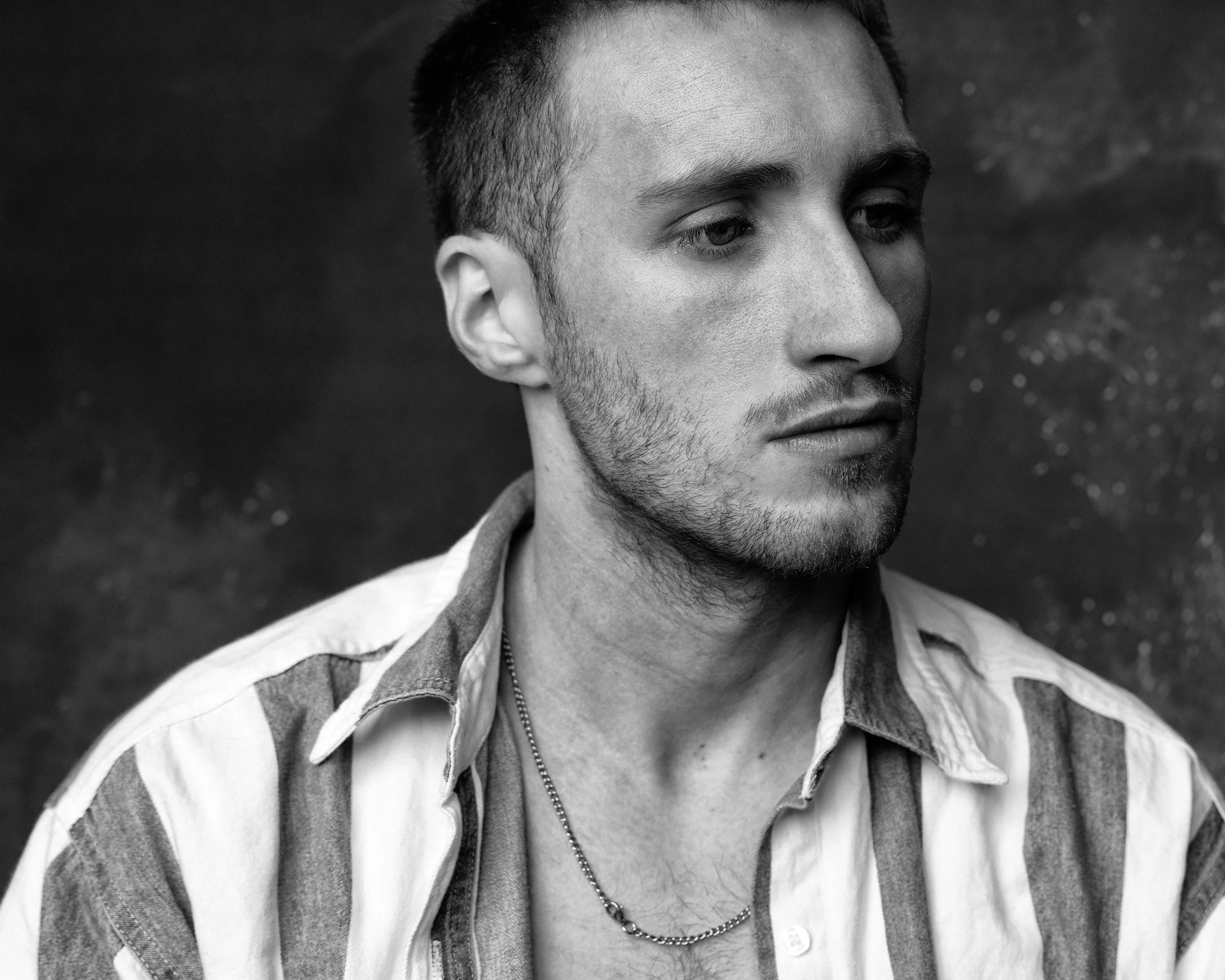 Close-up black and white photo of a young man's face, looking to the right, with short dark hair and a beard, wearing a collared shirt and a chain necklace.