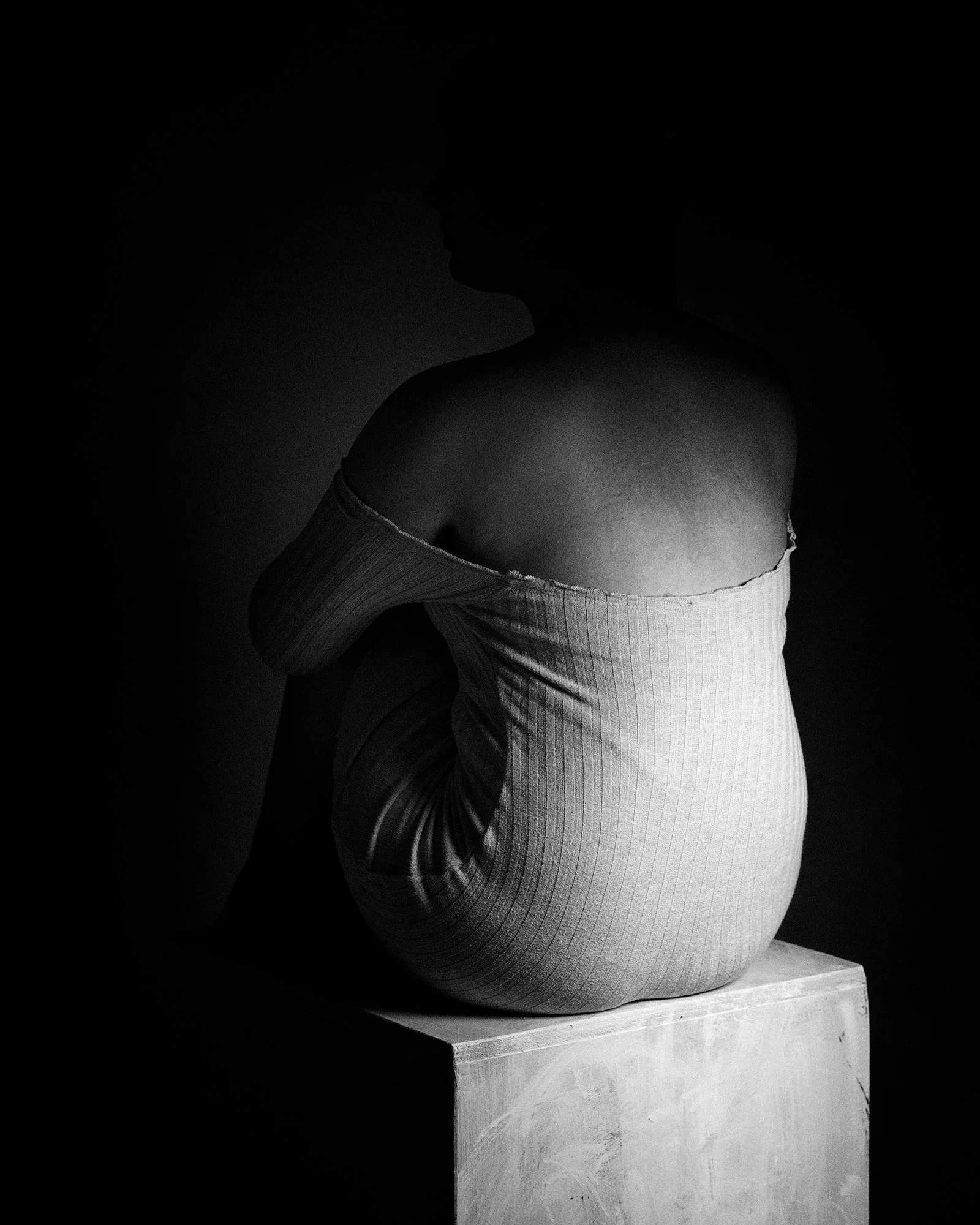 Black and white photo of a woman sitting on a block, wearing an off-the-shoulder top with her head turned away, creating a silhouette.