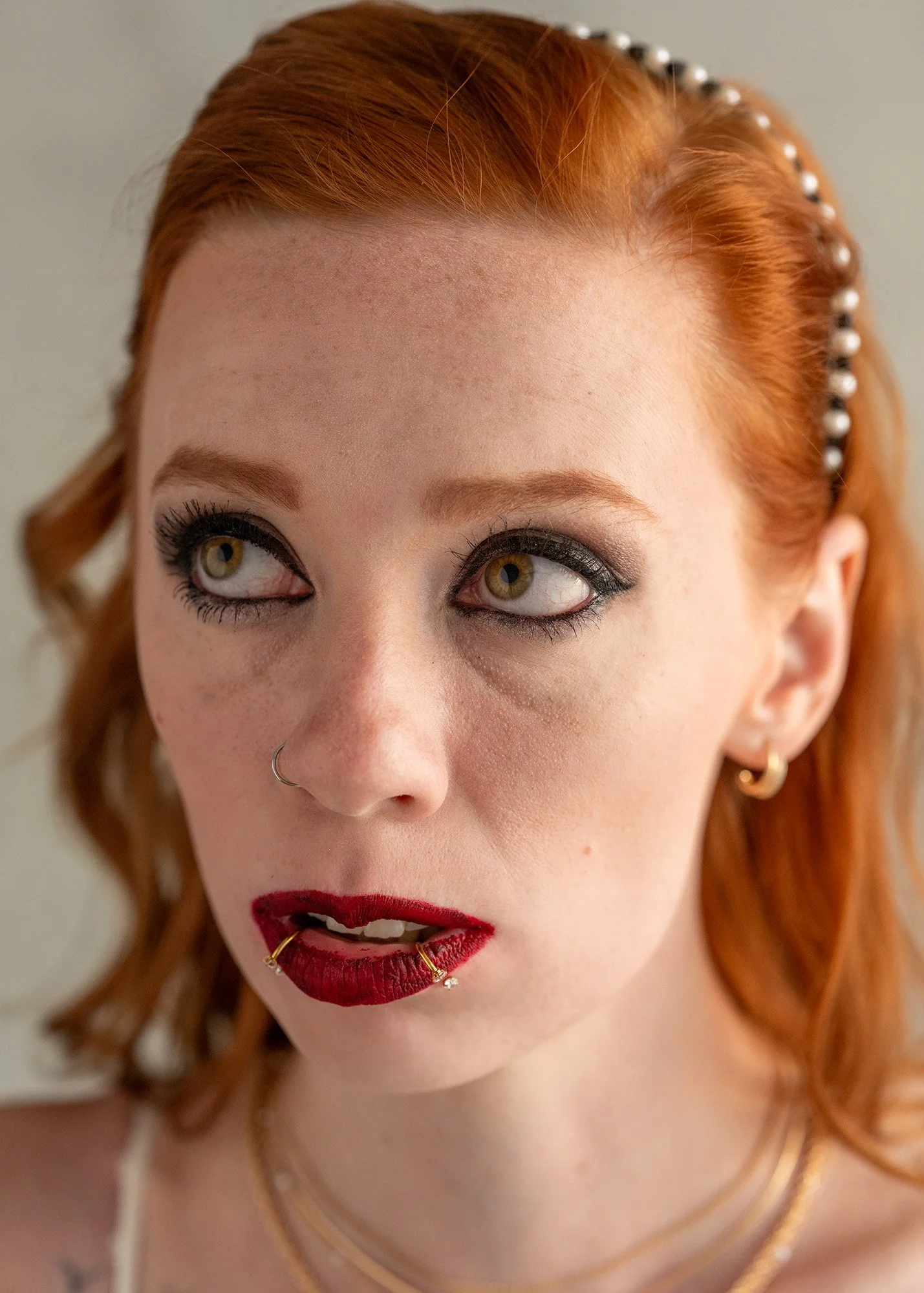 Close-up of a woman with red hair, makeup, piercings, and jewelry, looking upward and to the side.