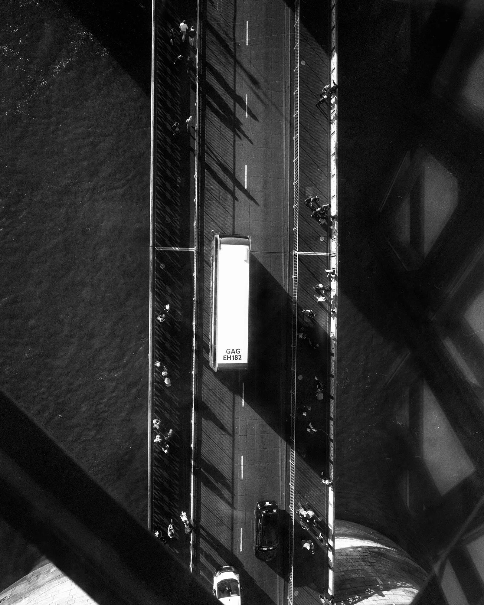 Bird's eye view of a city sidewalk with people walking, along with parked cars and a large bus, captured from above in black and white.