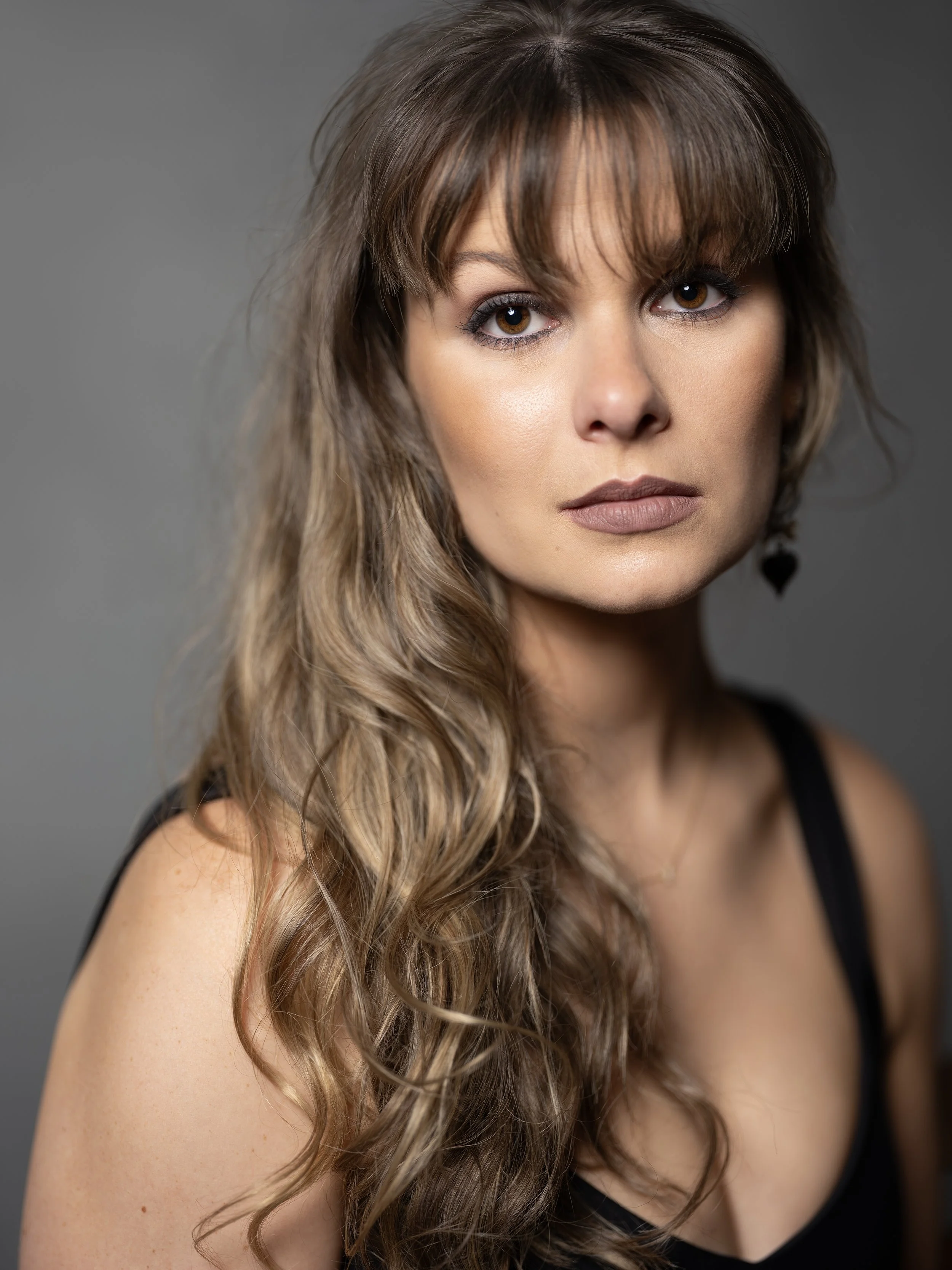 A woman with long wavy brown hair, wearing makeup, a black sleeveless top, and earrings, posing against a gray background.
