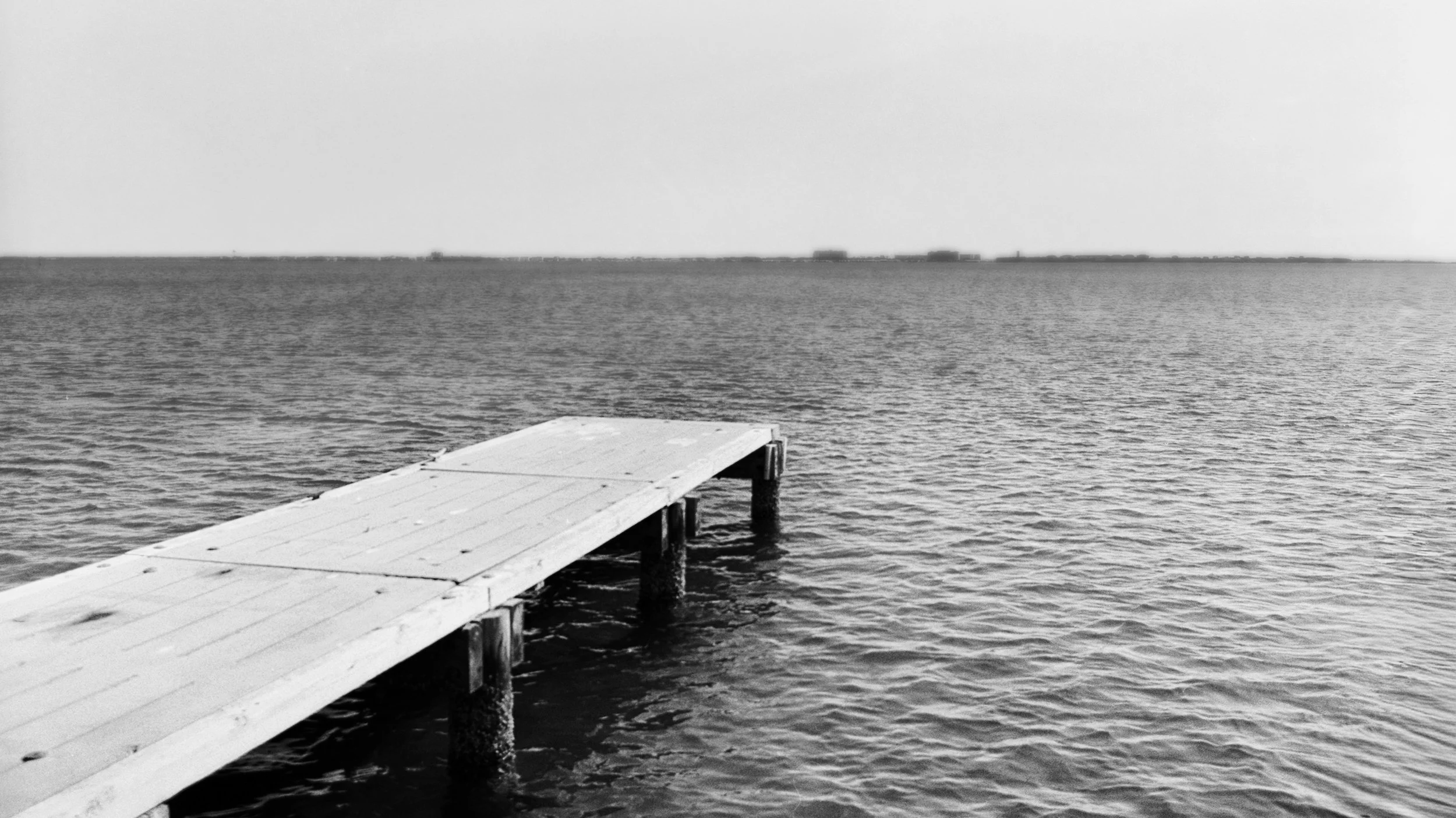 A black and white photo of a wooden dock extending into a calm body of water with a distant horizon.