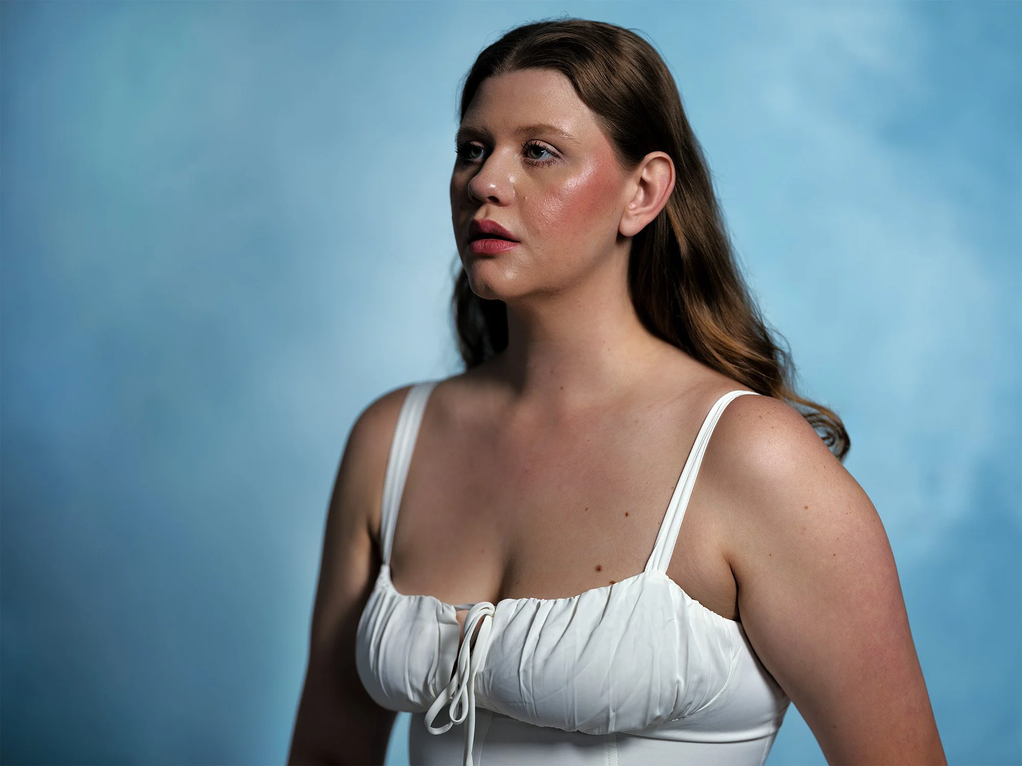 A woman with long brown hair and wearing a white top stands against a blue background, looking to the side.