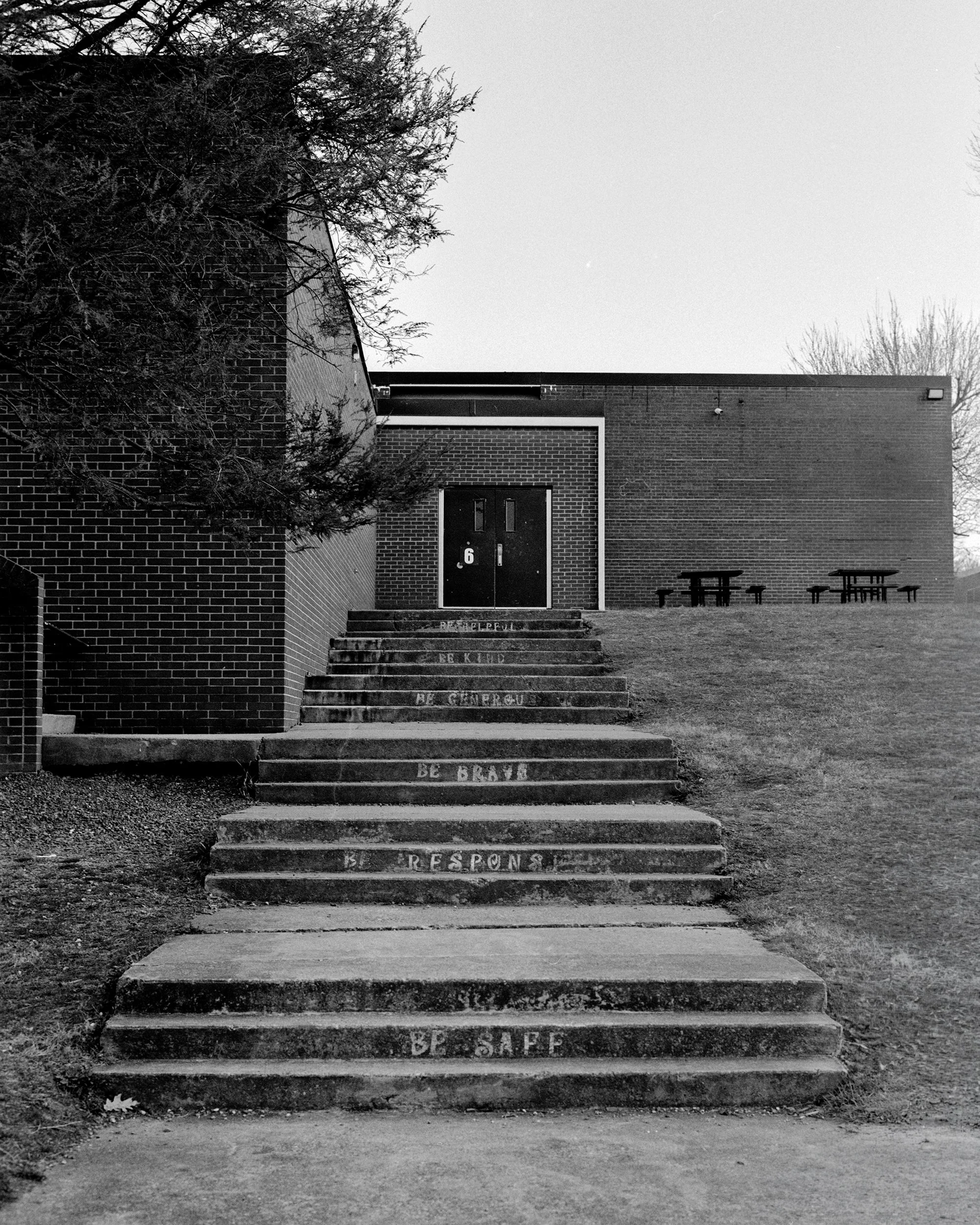 Black and white photograph of building entrance with stairs engraved with words like "Respect" and "Be Brave." There are two benches on the right side and a tree on the left side of the image.