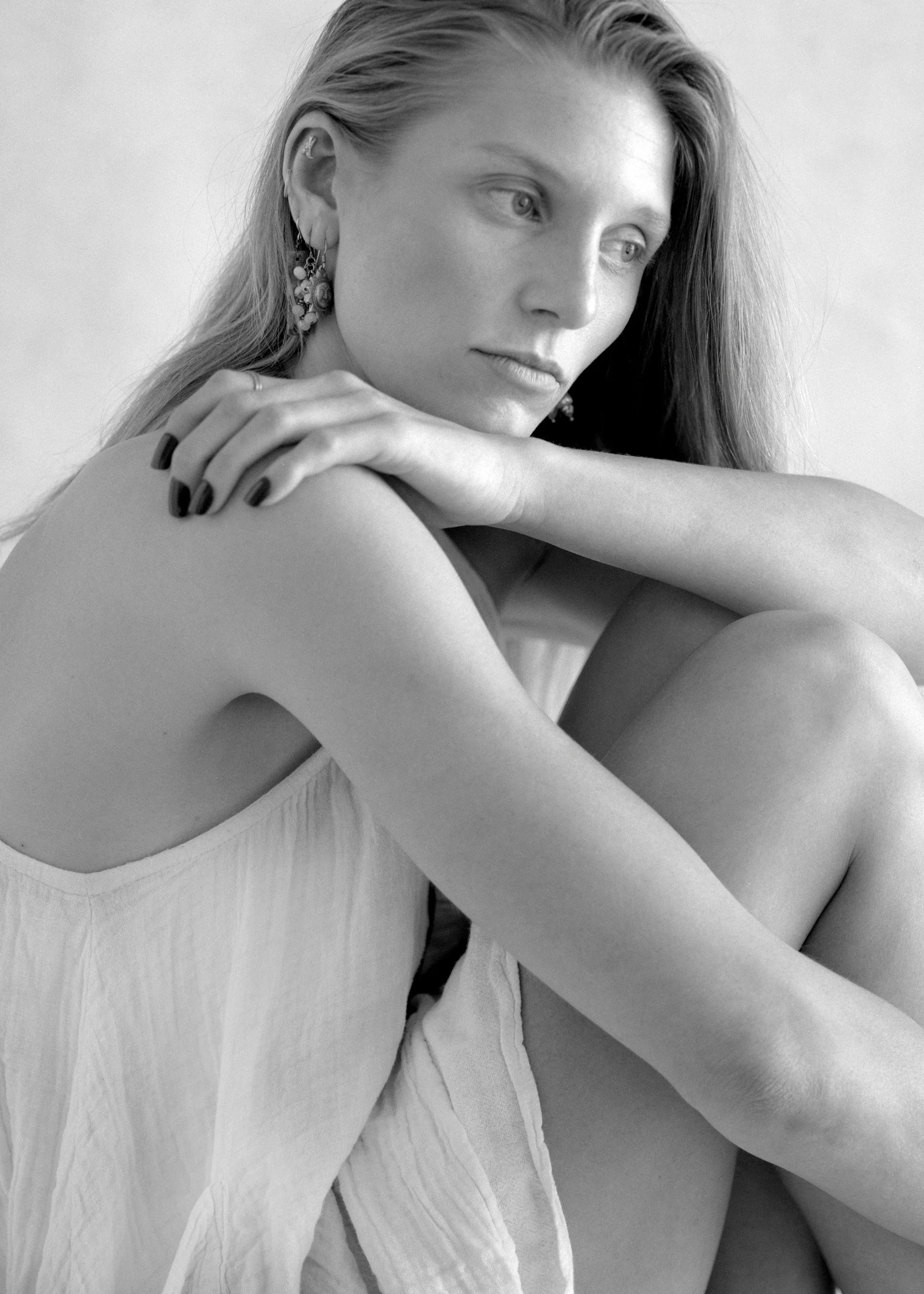 Black-and-white photo of a woman with long hair, wearing a sleeveless top and earrings, resting her arms on her knees and gazing thoughtfully.