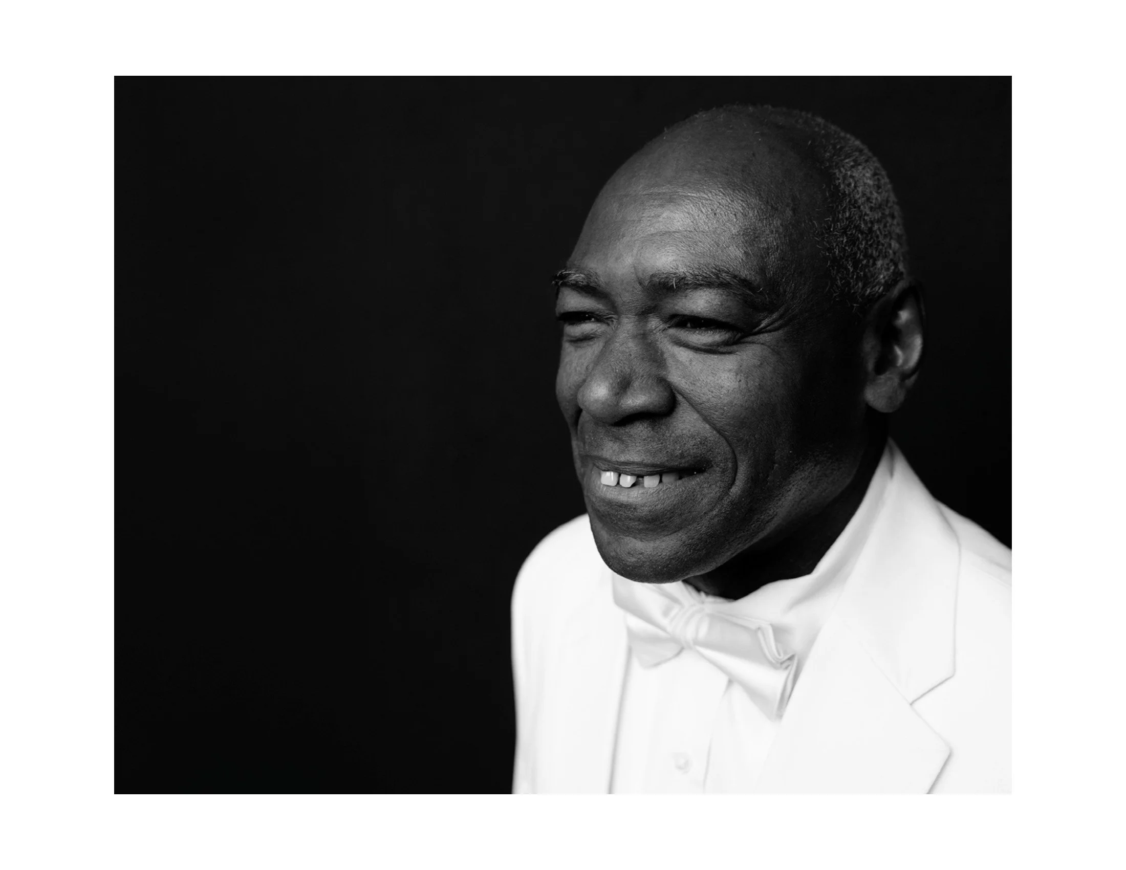 Close-up portrait of a smiling black man wearing a tuxedo with a white bow tie against a black background.