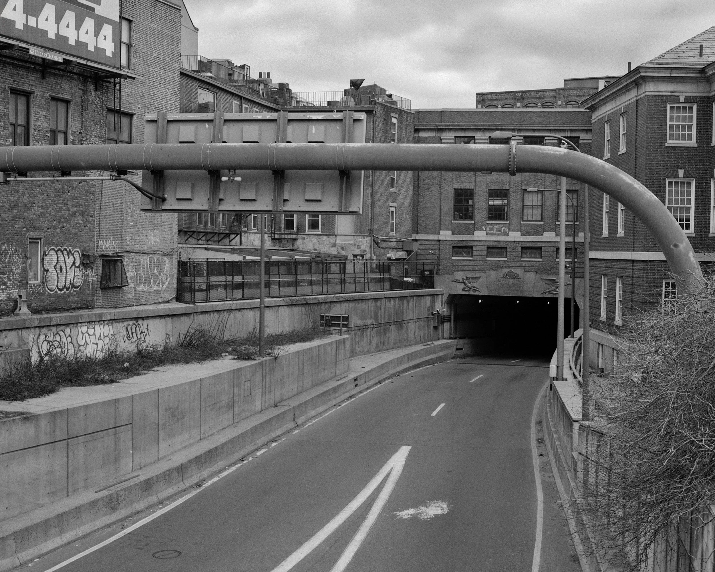 A city street scene captured in black and white, showing a curved road with a street side barrier, brick apartment buildings with multiple windows, graffiti on the lower part of a building wall, a large pipe overhead, and a cloudy sky.