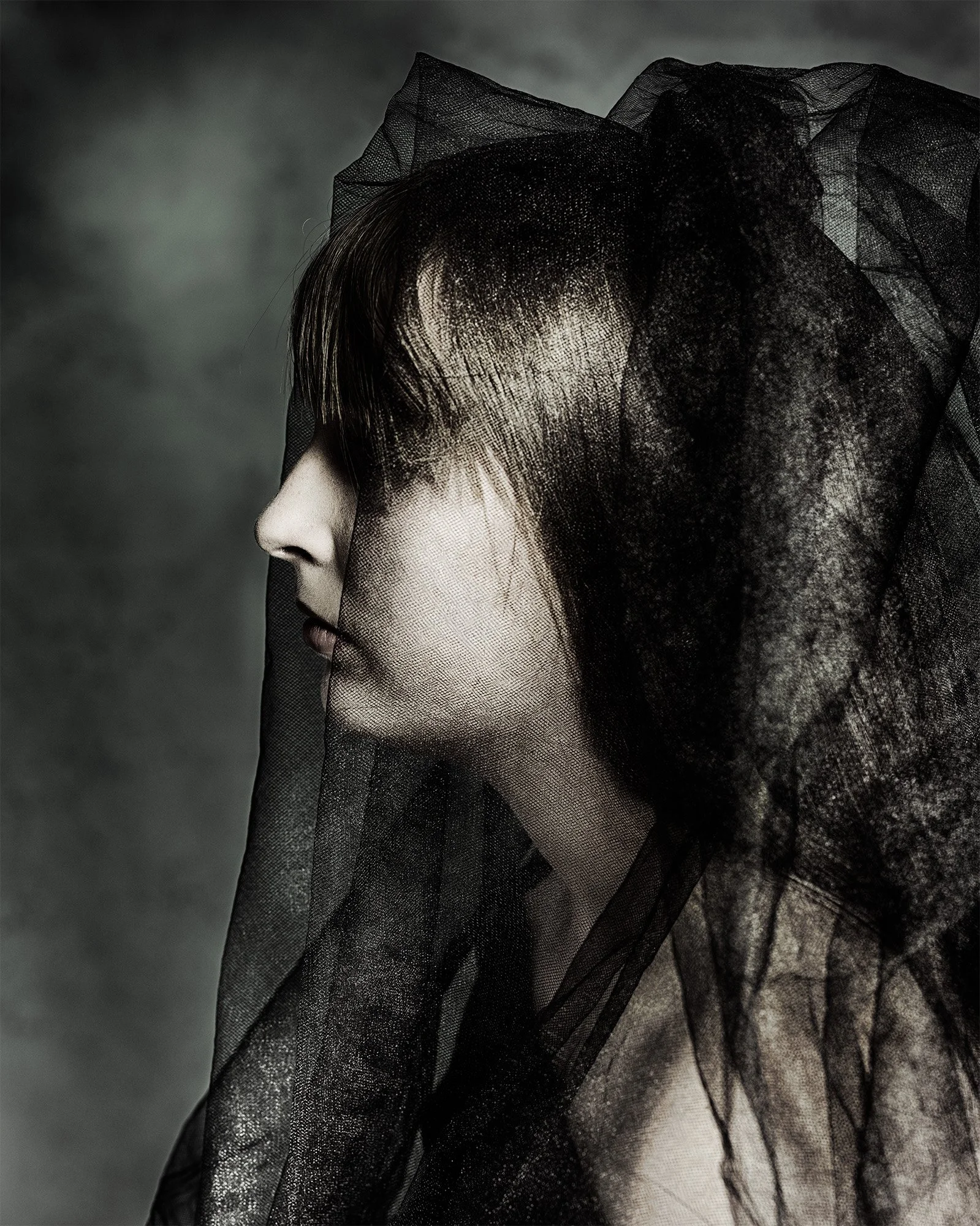 Side profile of a woman with black tulle fabric covering her head and shoulders, with a dark background.
