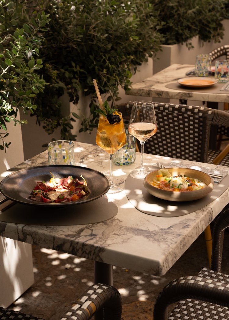 Lunch on the terrace of Chez Junior: a marble table with two plates, a spritz and a glass of white wine, in the dappled shade of the olive trees