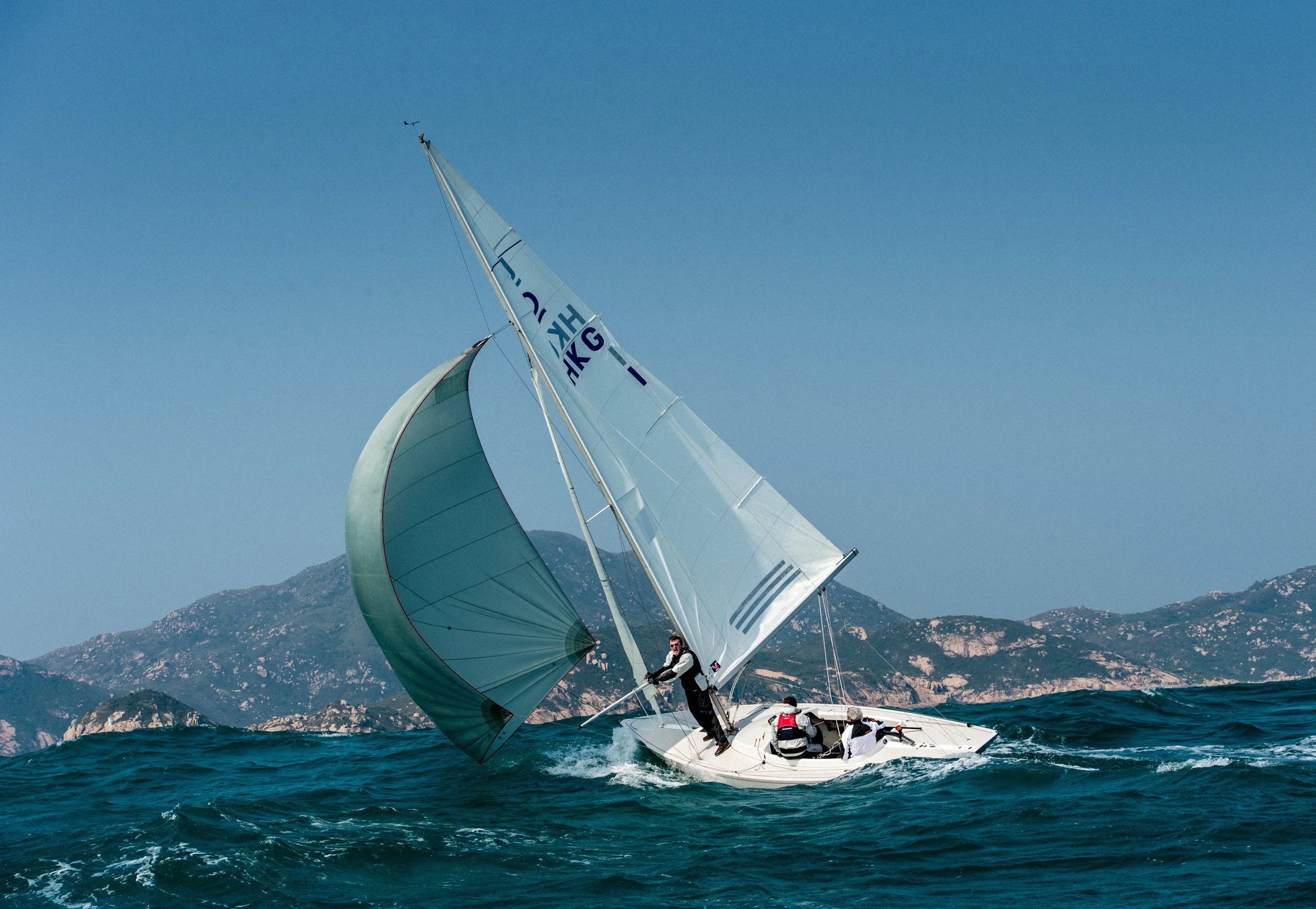 Sailboat with spinnaker sail catching wind, sailing on choppy water near mountainous coastline, with three crew members on board.