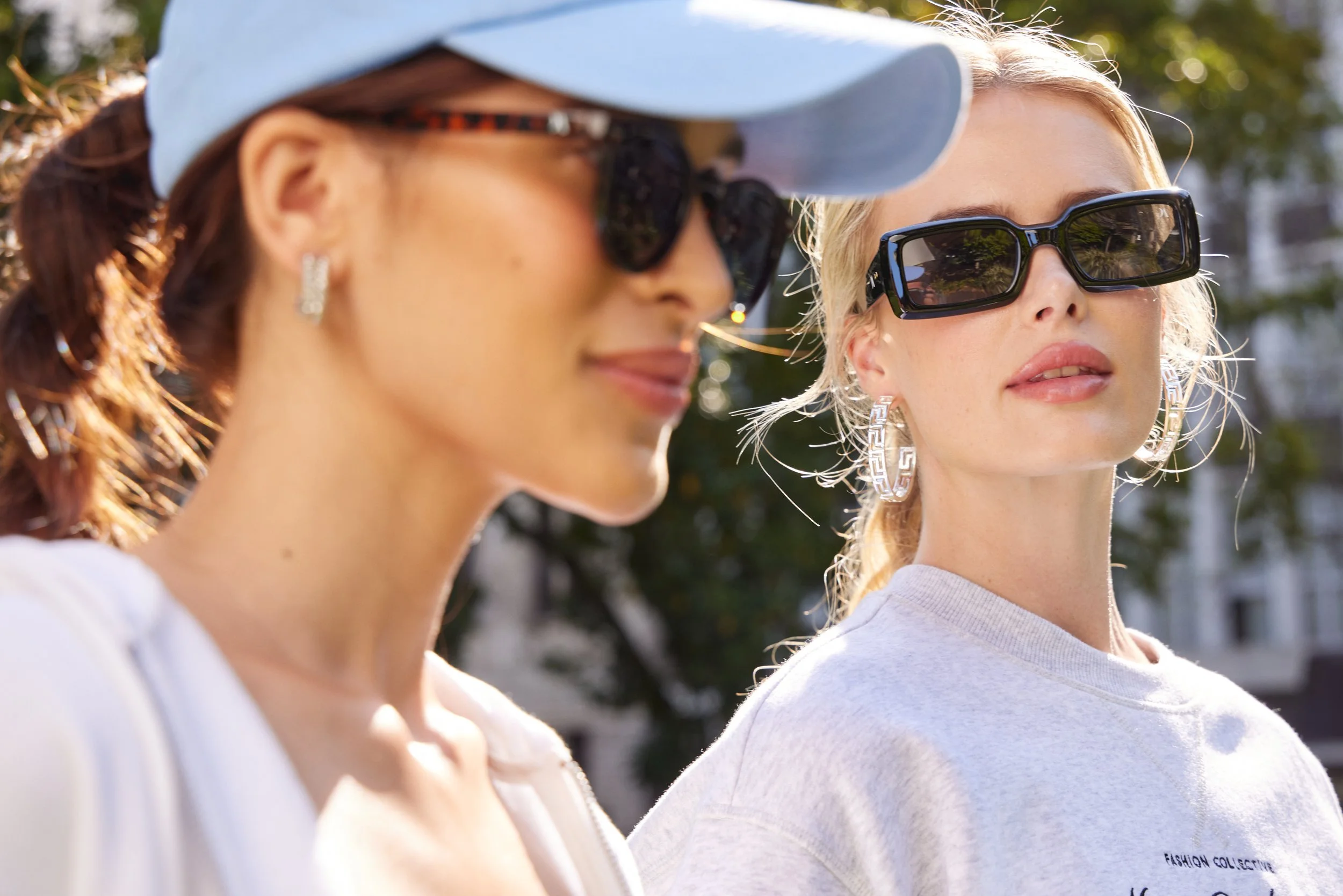 Two women wearing sunglasses, one with a white baseball cap, outdoors with sunlight filtering through trees.
