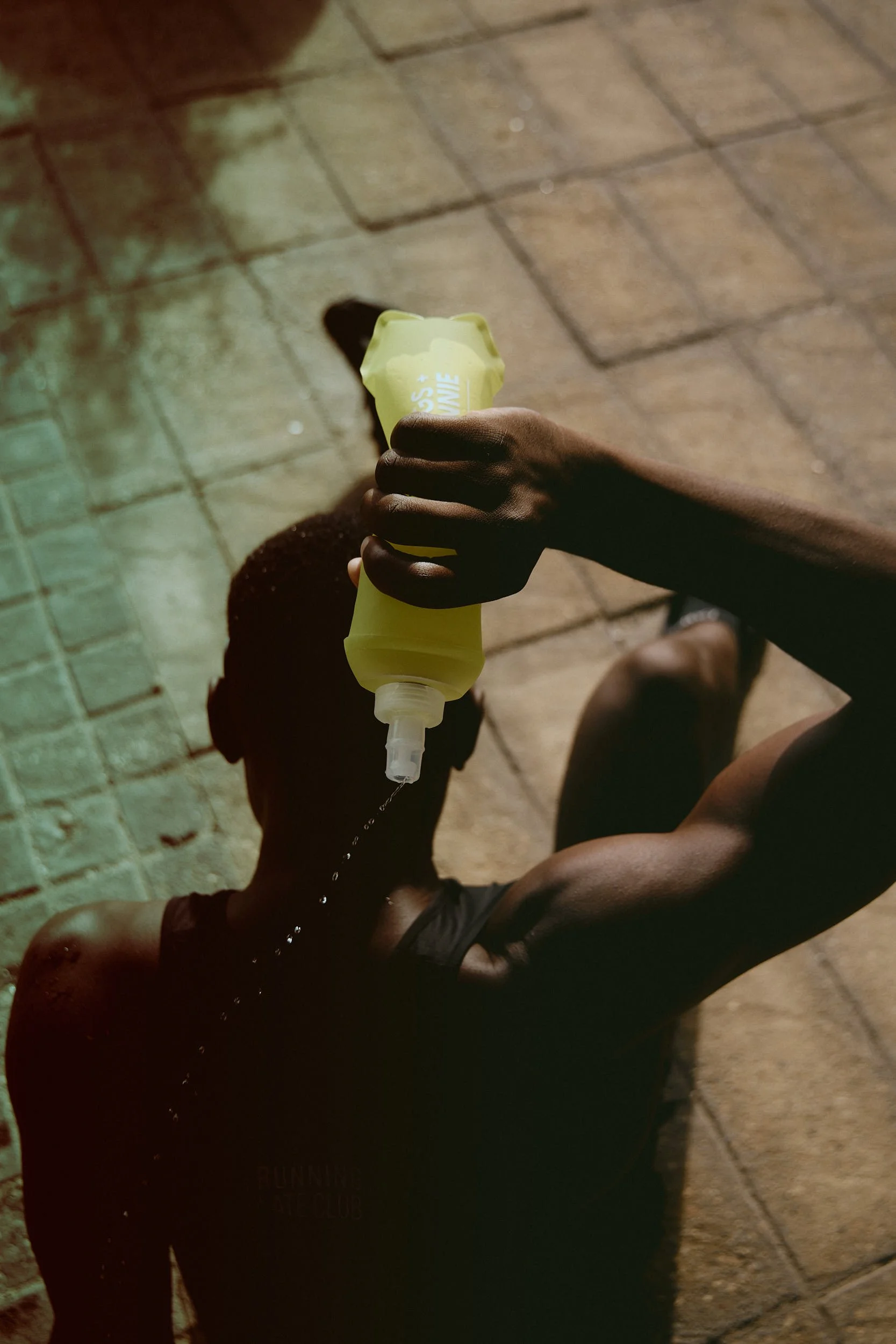 A person with dark skin is sitting on the ground and holding a yellow hand sanitizer bottle, squeezing it to dispense sanitizer onto their hand. They are wearing dark clothing and a dark hat, and the ground is made of brick or tile.