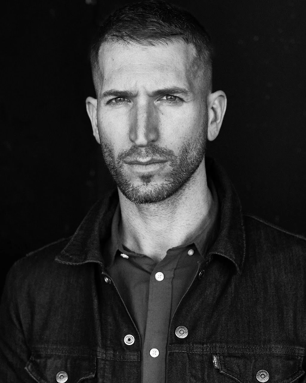 Black and white close-up portrait of a man with short hair and beard, wearing a denim jacket over a button-up shirt, against a dark background.
