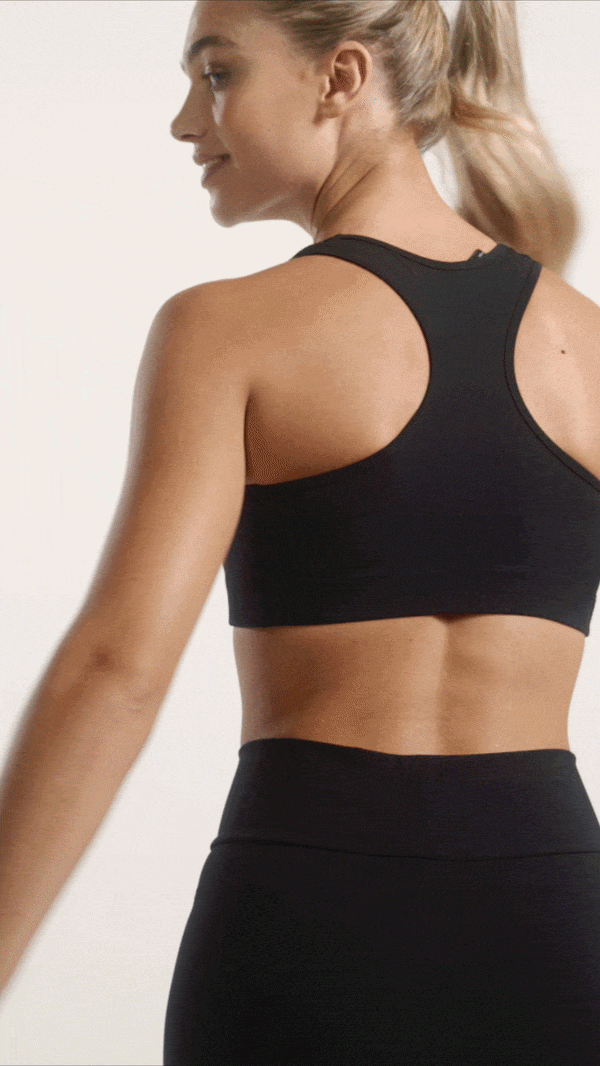 A woman wearing a black sports bra and black leggings, looking to the side, with her hair in a ponytail, against a plain white background.