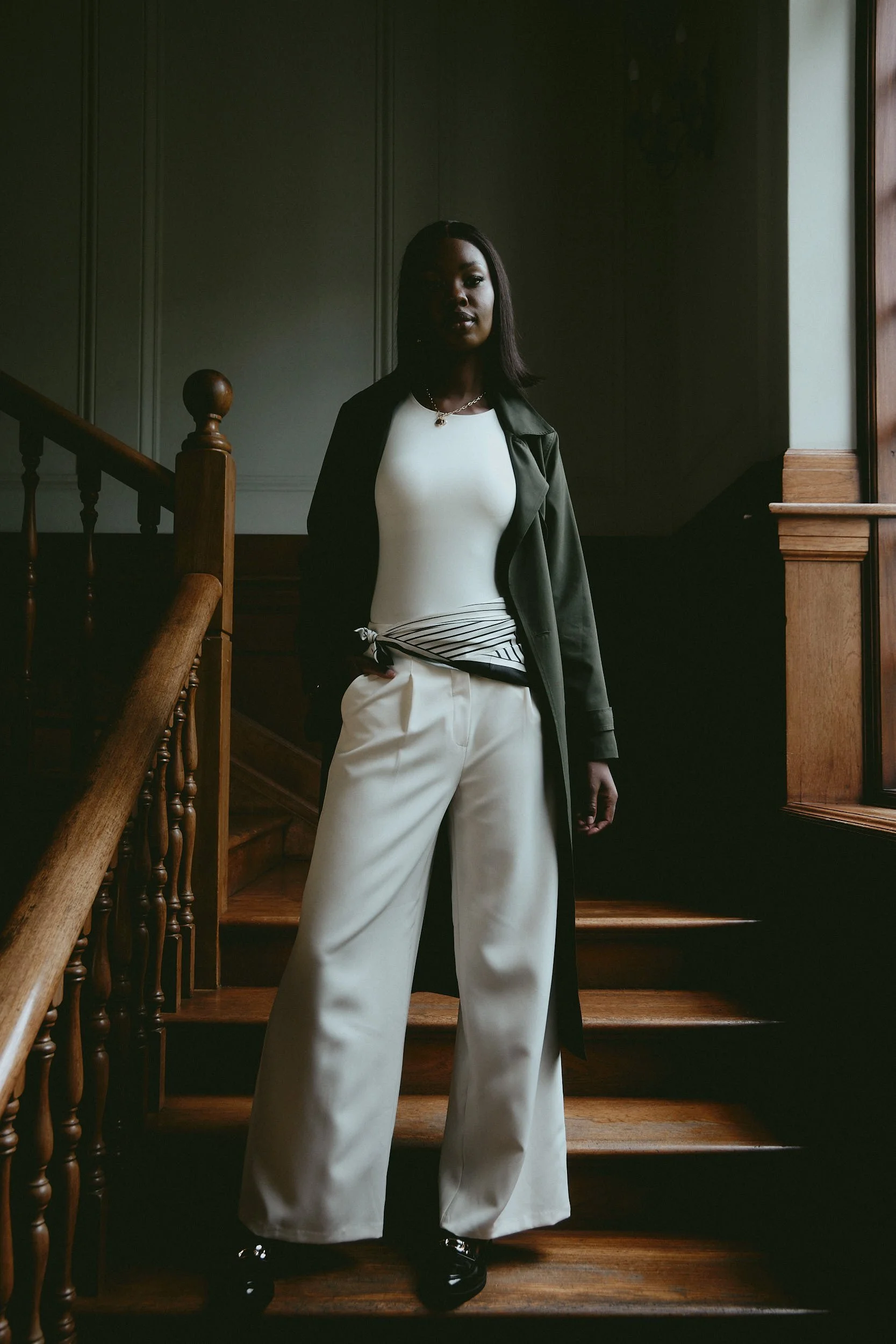 A woman with dark hair wearing a white top, wide-legged white pants, a dark green coat, black shoes, a necklace, stands on a wooden staircase inside a house with gray panel walls and natural light coming through a window.