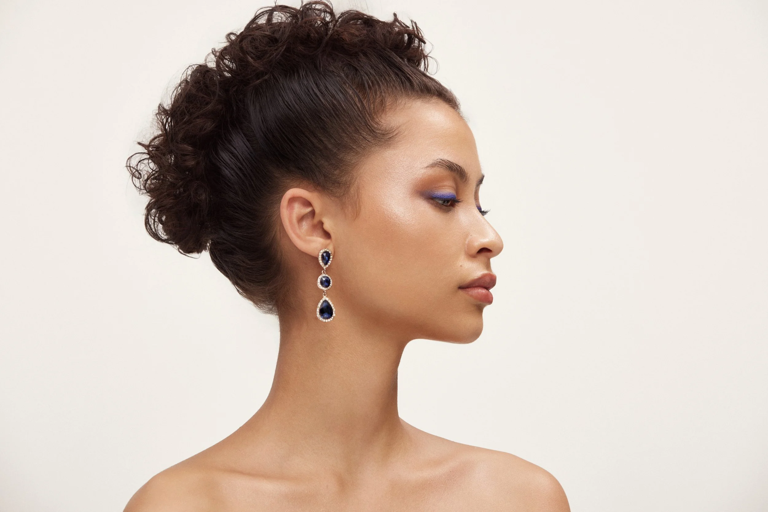 A side profile of a woman with dark curly hair styled in an elegant updo, wearing large blue gemstone drop earrings, with makeup featuring purple eyeshadow, against a plain neutral background.