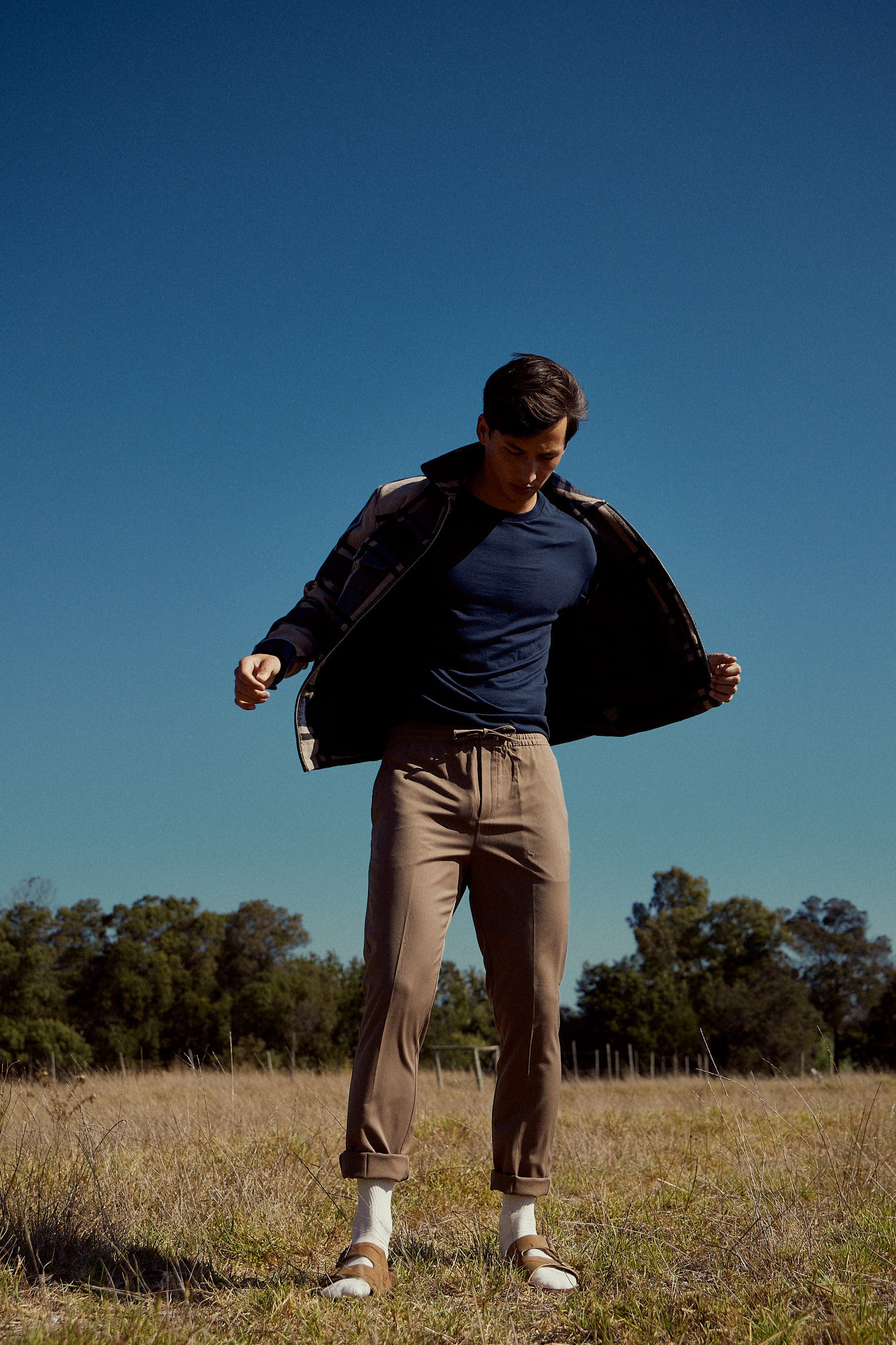 A young man wearing a navy blue t-shirt, beige pants, and a jacket is standing in a grassy field with trees in the background, looking downward, with a clear dark blue sky overhead.
