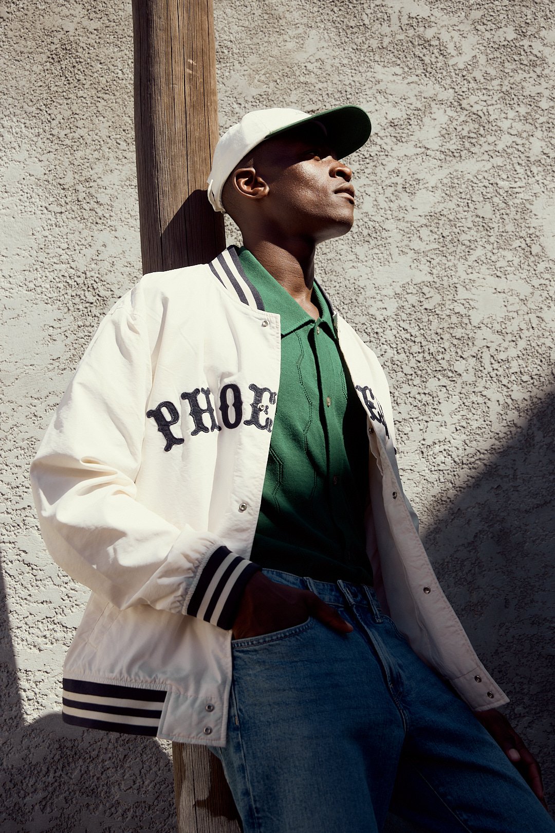 A young man leaning against a wooden pole with a textured wall in the background, wearing a white varsity jacket with black lettering, green polo shirt, baseball cap, and jeans, looking upward.
