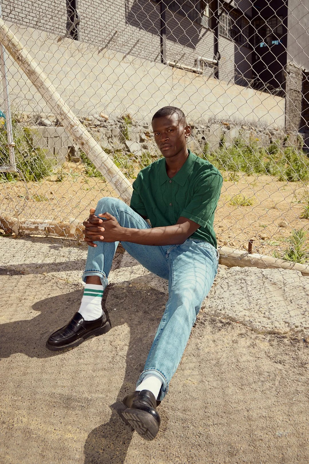 A young man sitting on the ground near a chain-link fence with a build up of soil and a building in the background.