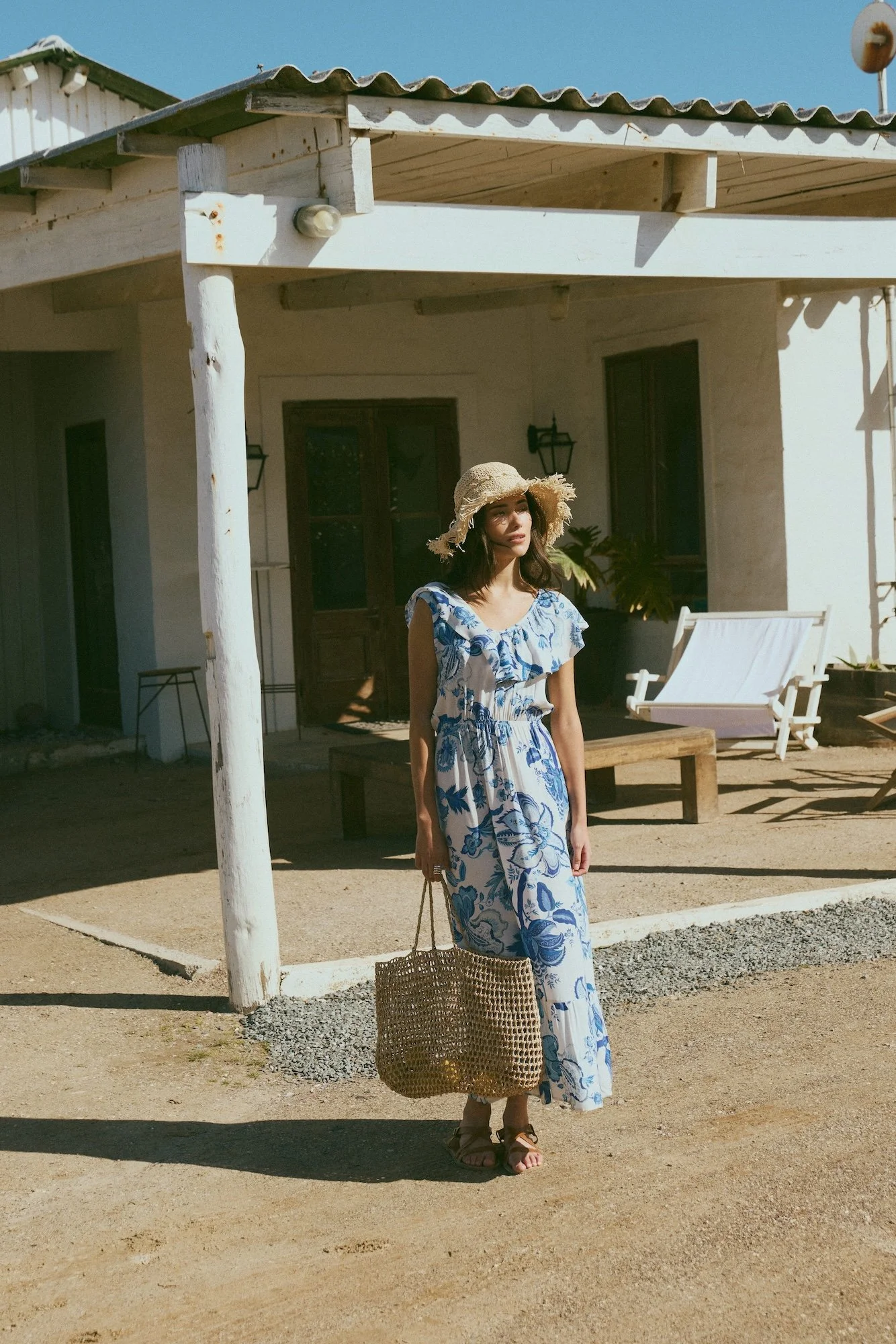 Woman in a blue and white floral dress with a wide-brim straw hat, holding a woven bag, standing outside a rustic house with a porch, wooden furniture, and patio chairs.