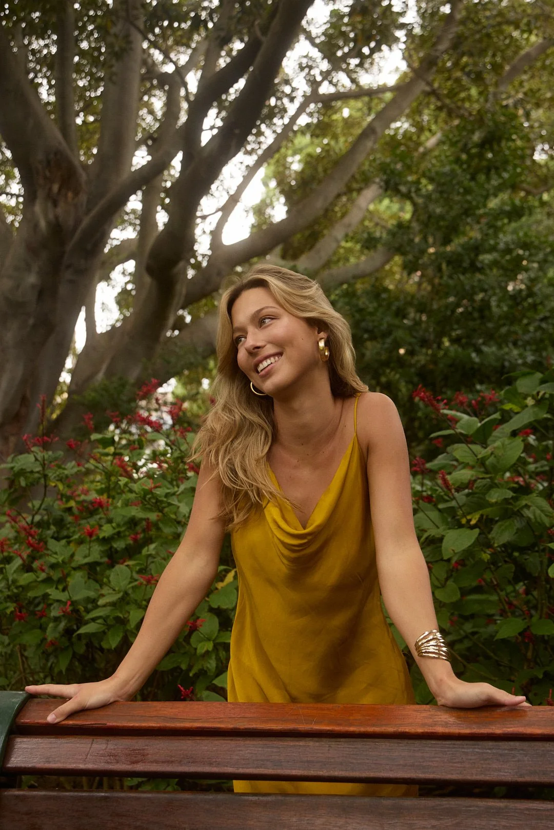 A woman with blonde hair wearing a yellow dress and jewelry, leaning on a wooden railing outdoors surrounded by greenery and flowers, smiling and looking to the side.