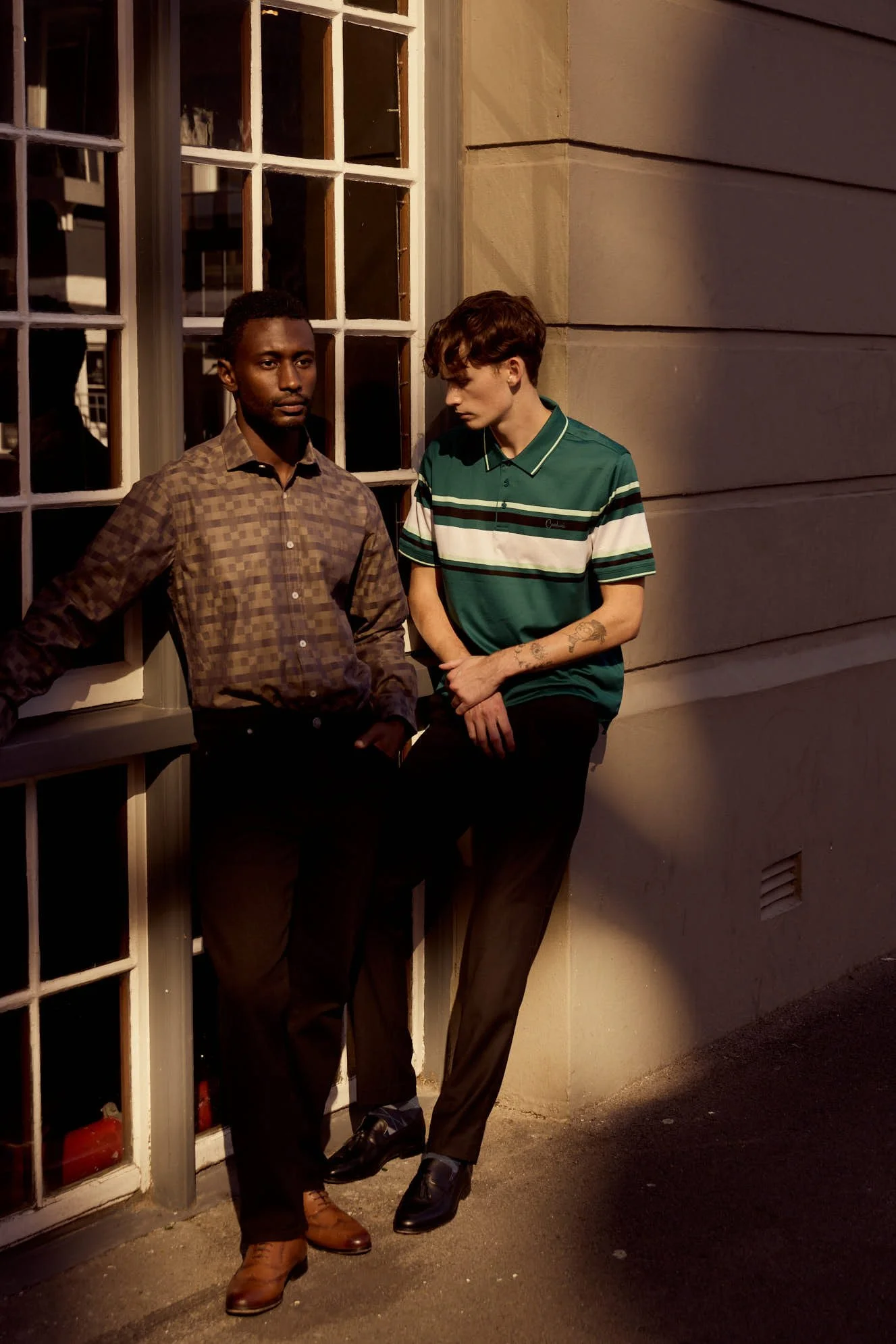 Two young men standing outside near a building with large windows, one looking at the camera and the other with head bowed, both dressed in casual but stylish clothing, with the scene lit by warm evening light.