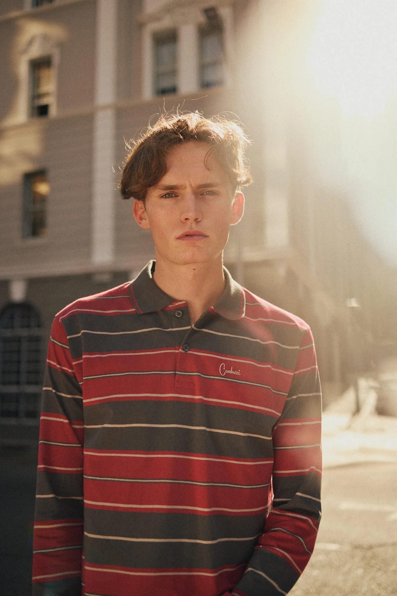 Young man with wavy brown hair wearing a red, gray, and black striped polo shirt standing outdoors in front of a building with a neutral expression.