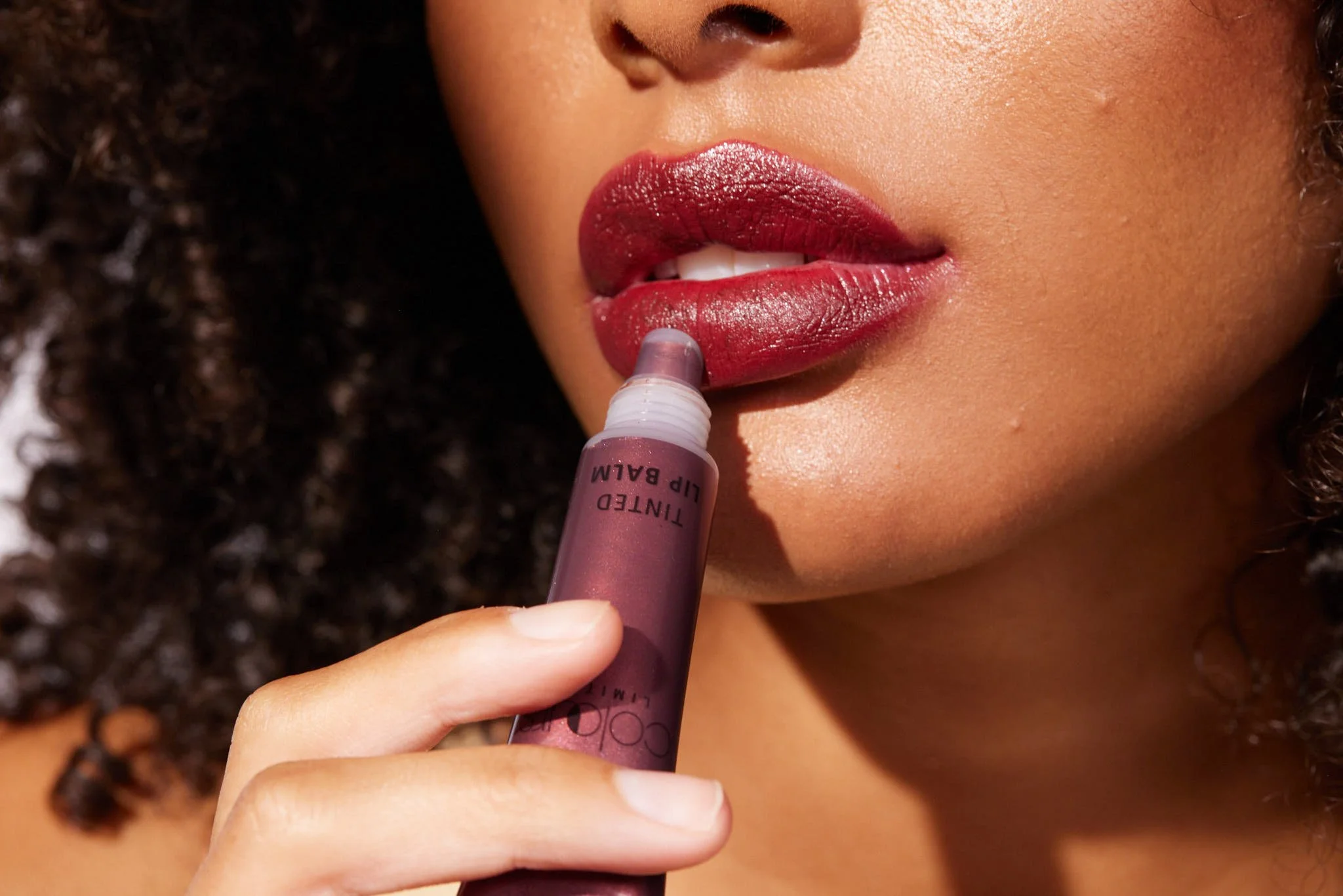 Close-up beauty portrait applying red lipstick, with a tinted lip balm tube visible, photography by Justin Munitz.