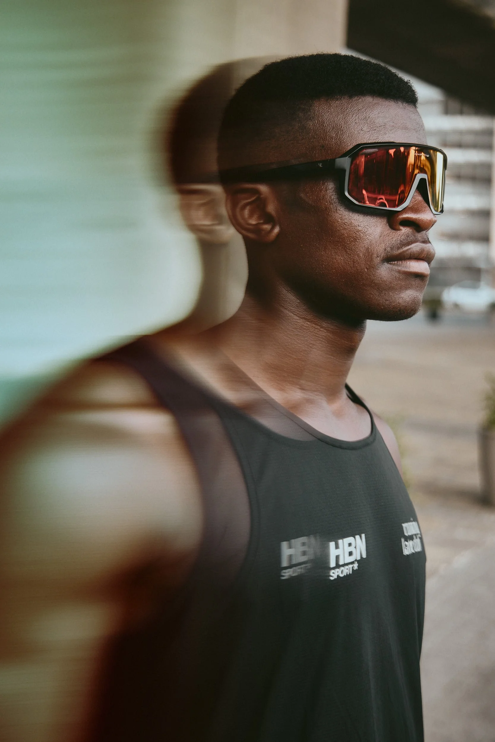 A young man wearing reflective sports sunglasses and a black tank top with white logos, standing outdoors during daytime.