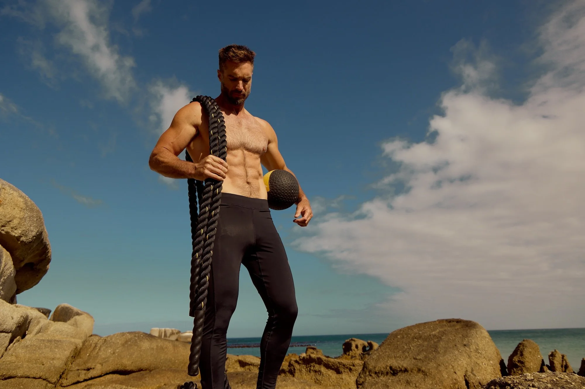 Active lifestyle photography by Justin Munitz — shirtless model holding a battle rope and medicine ball on a rocky beach