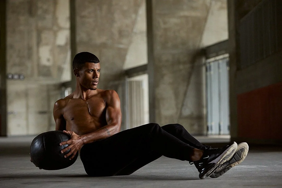 Active lifestyle photography by Justin Munitz - shirtless model seated on the floor holding a medicine ball, in a warehouse-style space.