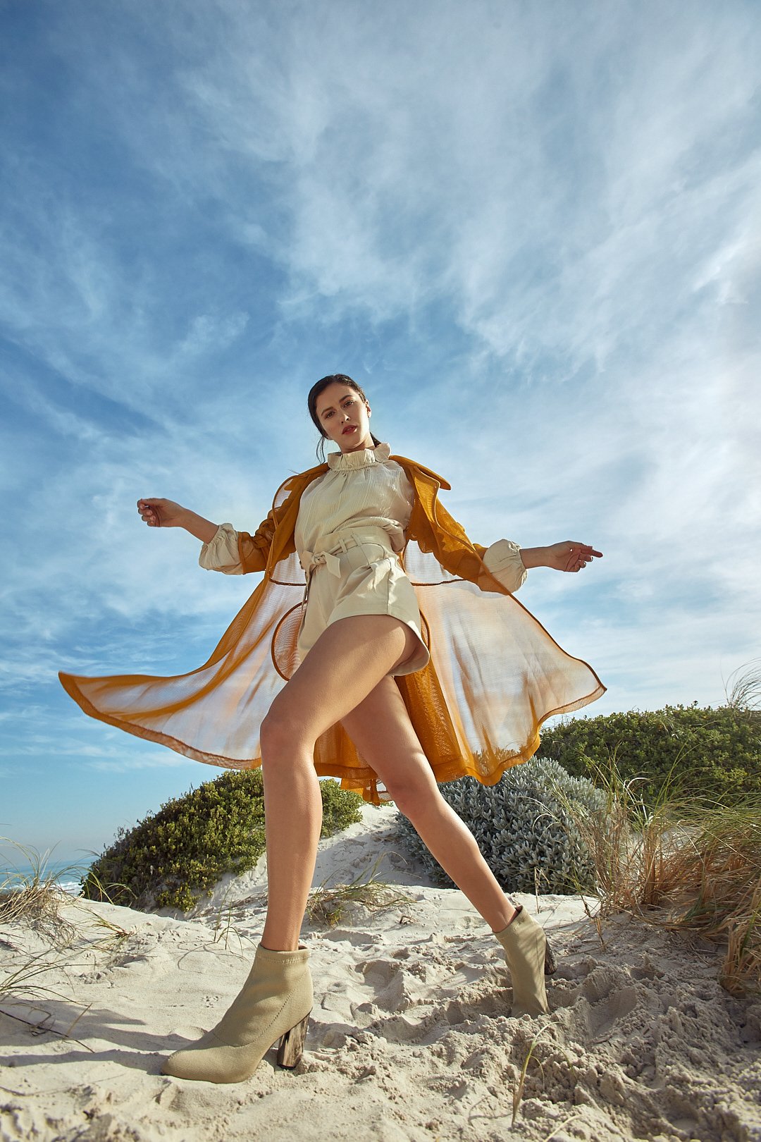Fashion photography by Justin Munitz . A woman standing on a sandy beach against a blue sky, wearing a beige outfit with an orange sheer coat, beige ankle boots, and posing with one leg raised.