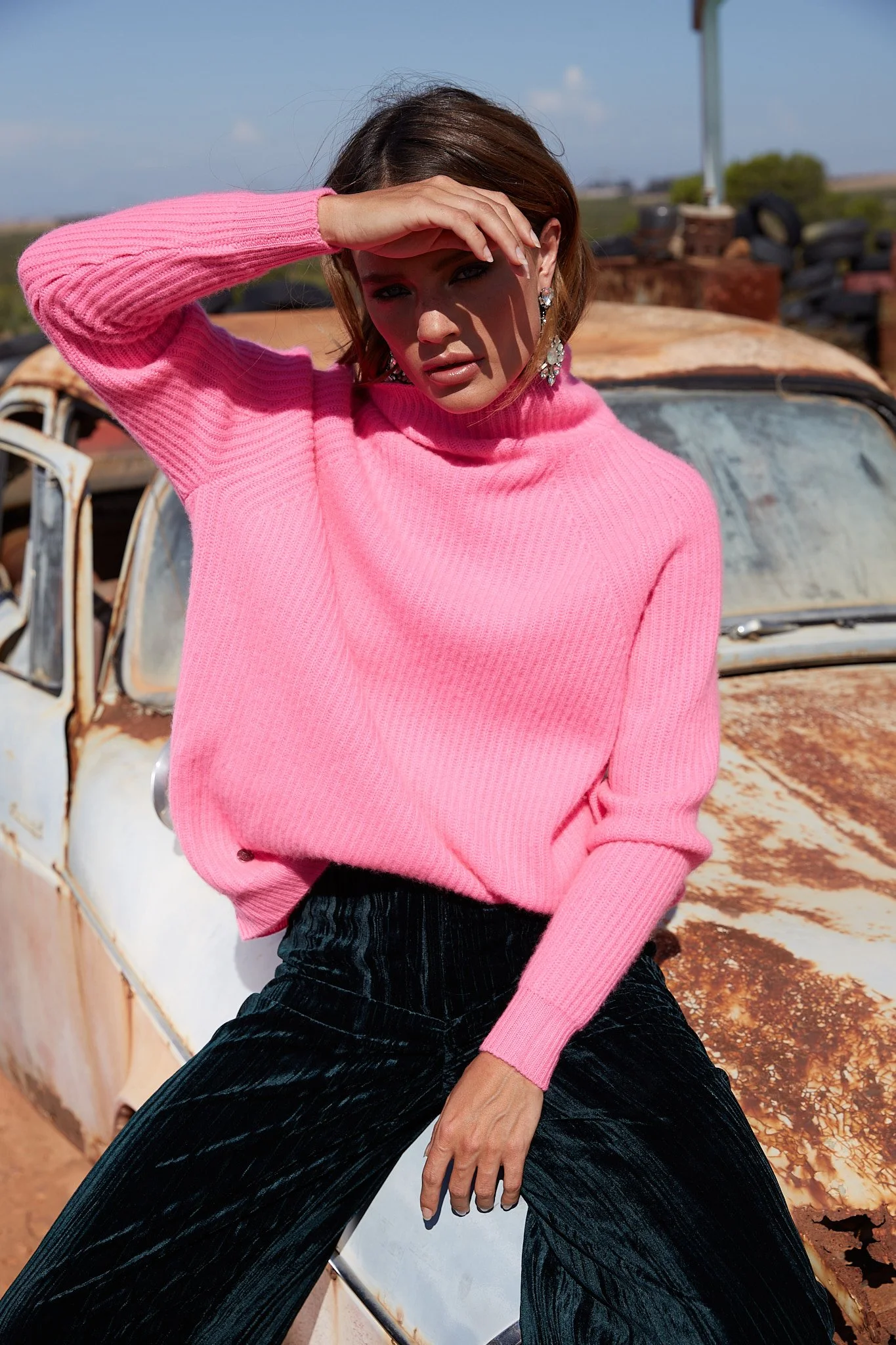 A woman with shoulder-length brown hair wearing a pink knit sweater and black pants, sitting on a rusted vintage car in an outdoor setting with tires and a pole visible in the background.