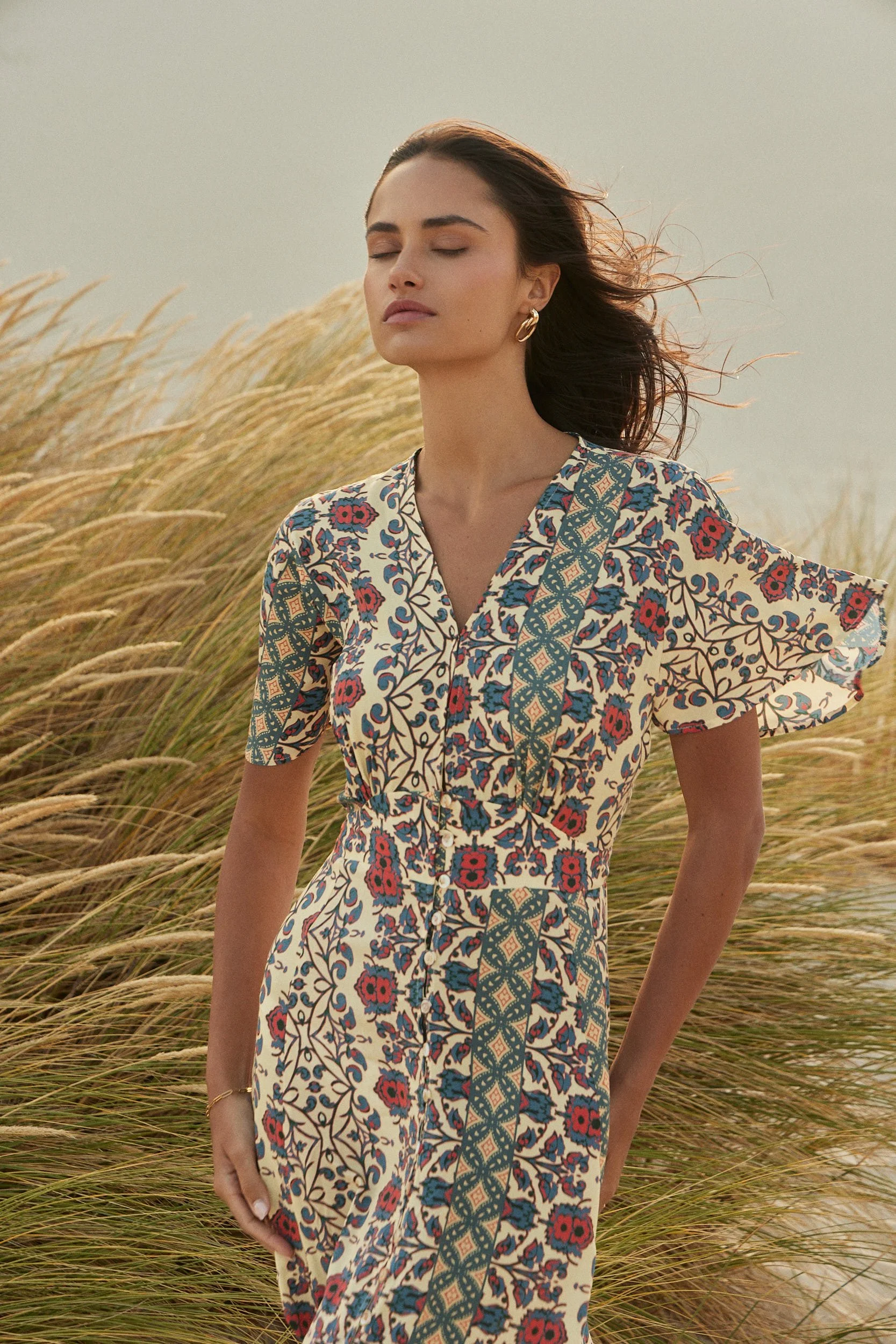 A woman with closed eyes and flowing hair standing in a grassy field during sunset, wearing a colorful patterned dress with earrings.