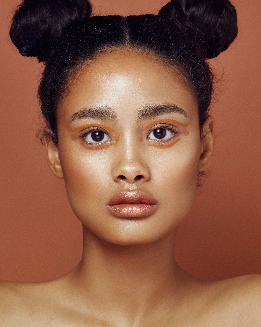 Beauty portrait with hair styled in two buns, natural makeup, and orange eyeliner accents, photography by Justin Munitz.