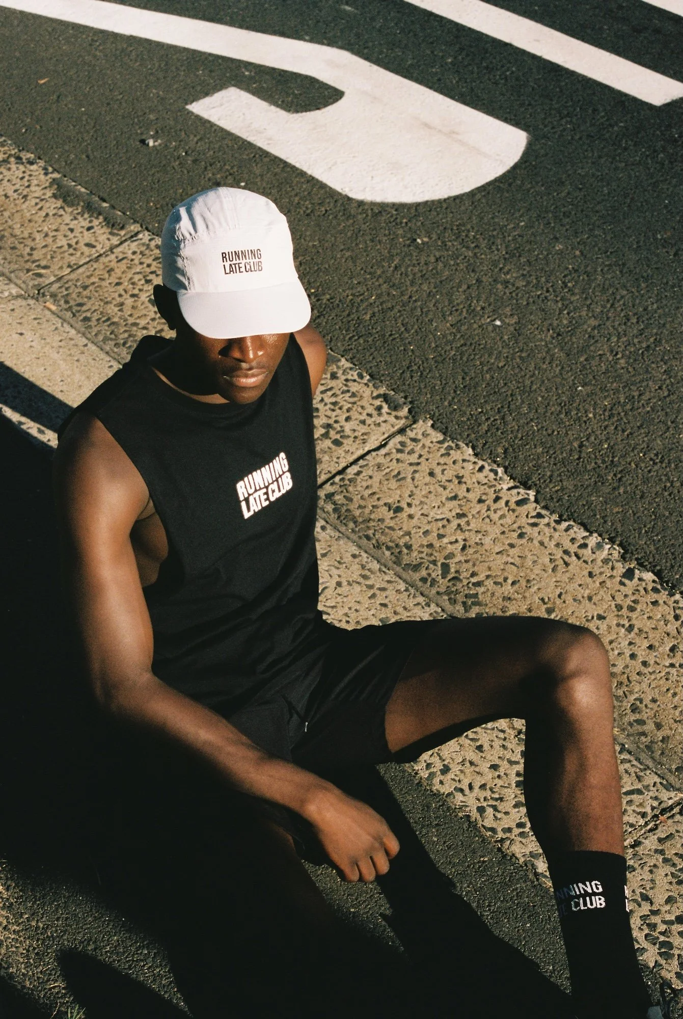 A person sitting on the sidewalk near a street curb, wearing a white cap and black sleeveless shirt with the text 'Running Late Club', with a painted white arrow and part of the word 'STOP' visible on the street.