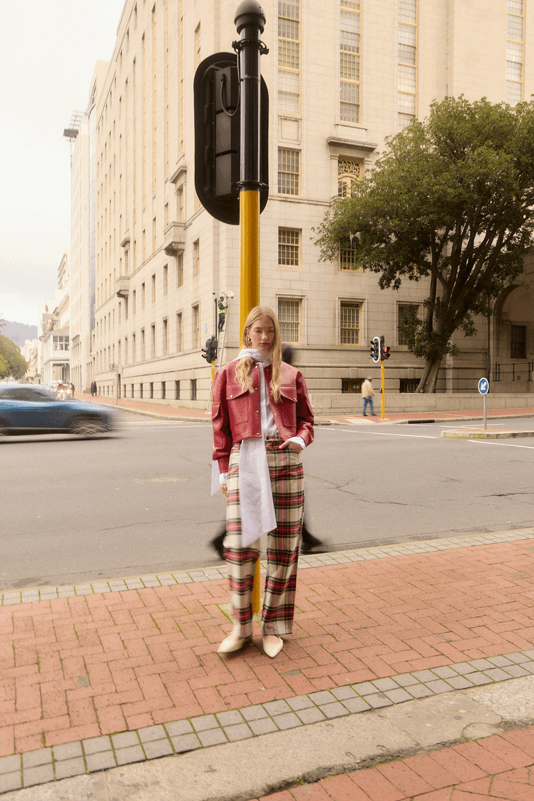 A young woman standing at a city street corner during daytime, wearing a red jacket, plaid pants, and cream-colored boots. She is leaning against a yellow pole with a black sign attached, with tall buildings and traffic in the background.