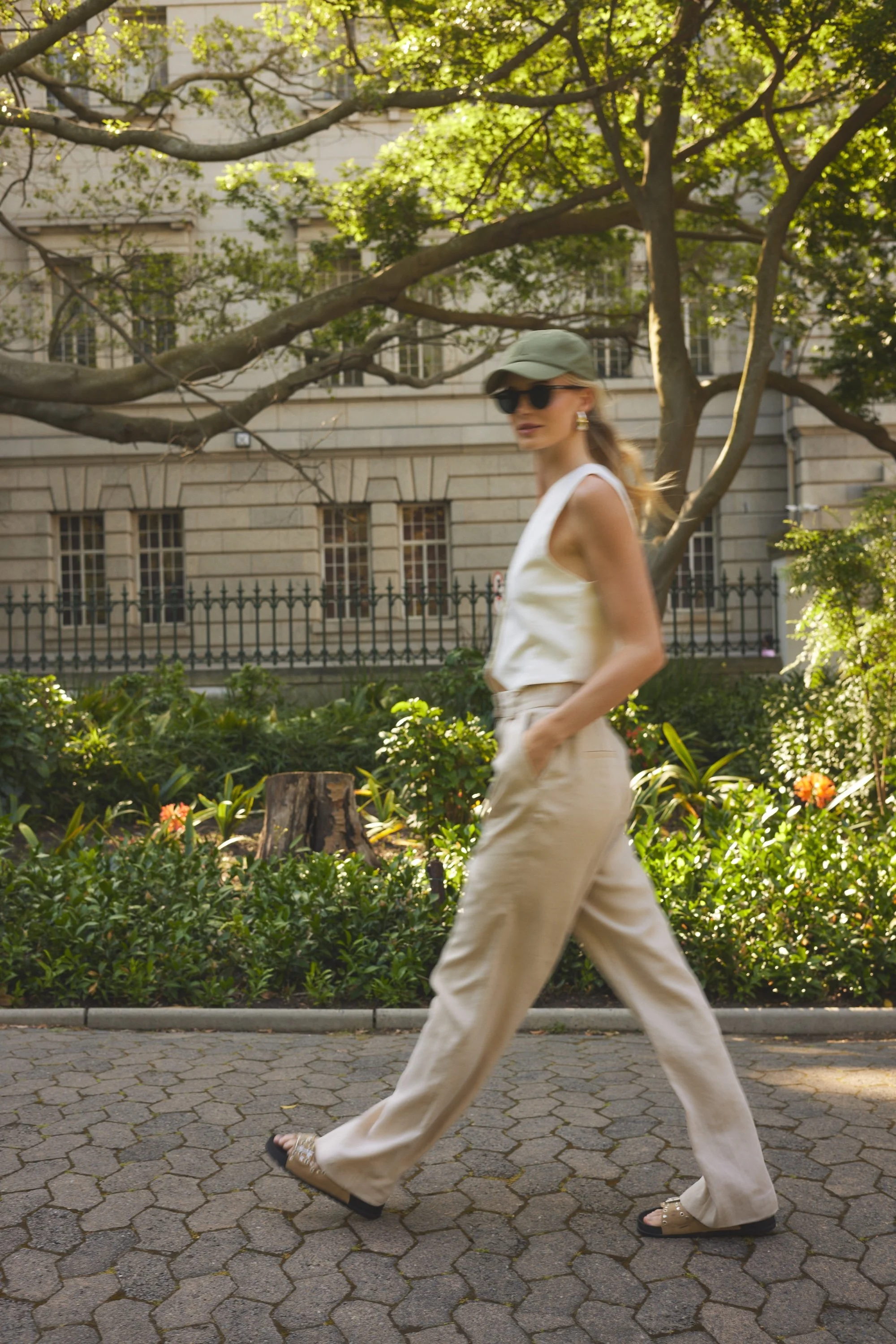 A woman walking outdoors on a sidewalk with a building and trees in the background. She is wearing a green cap, sunglasses, large earrings, a white sleeveless top, and beige wide-leg pants.