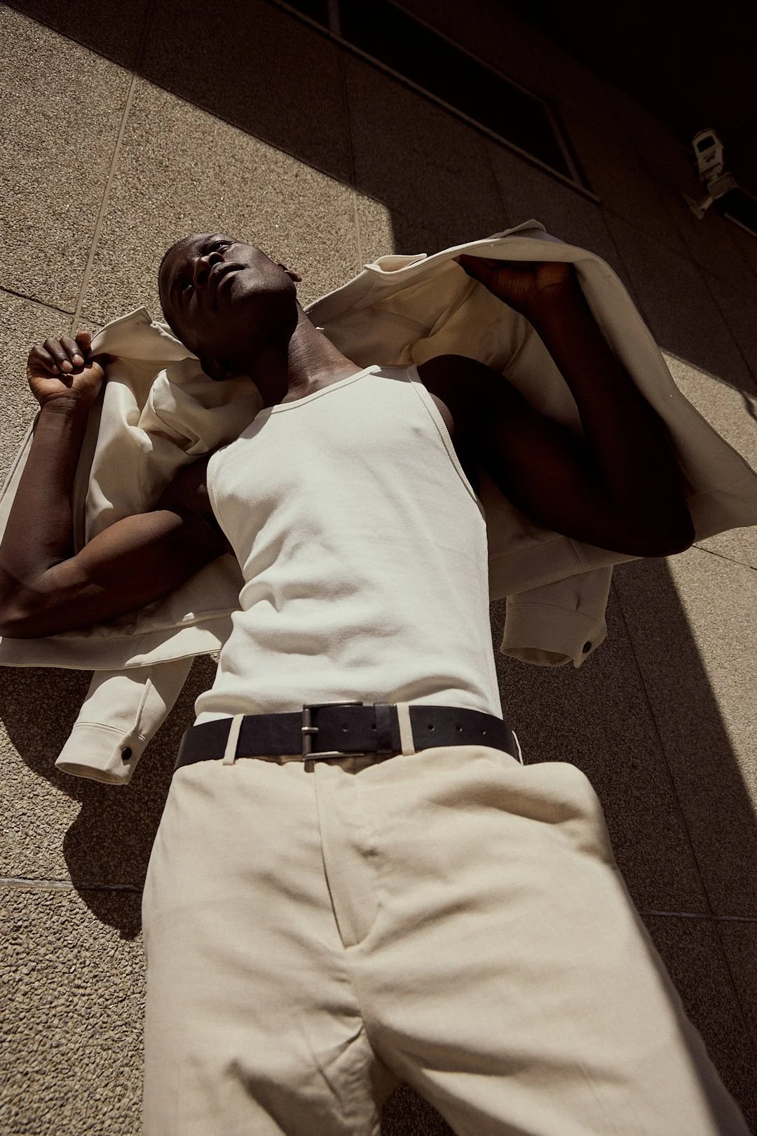 A man lying on the sidewalk with his eyes closed, wearing a white tank top and beige pants, with a beige jacket draped over him.