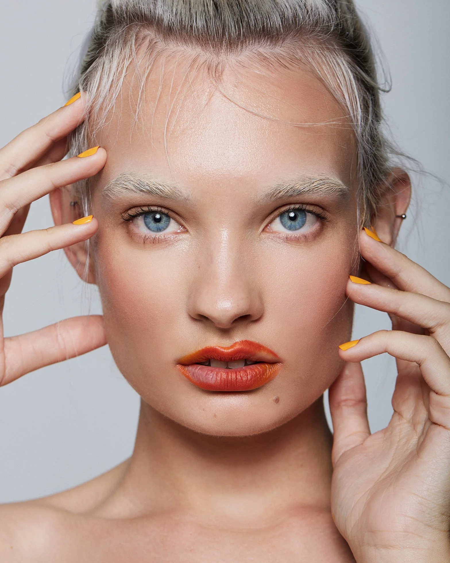 Close-up beauty portrait with orange lipstick and yellow-painted nails, touching her face with both hands, photography by Justin Munitz.