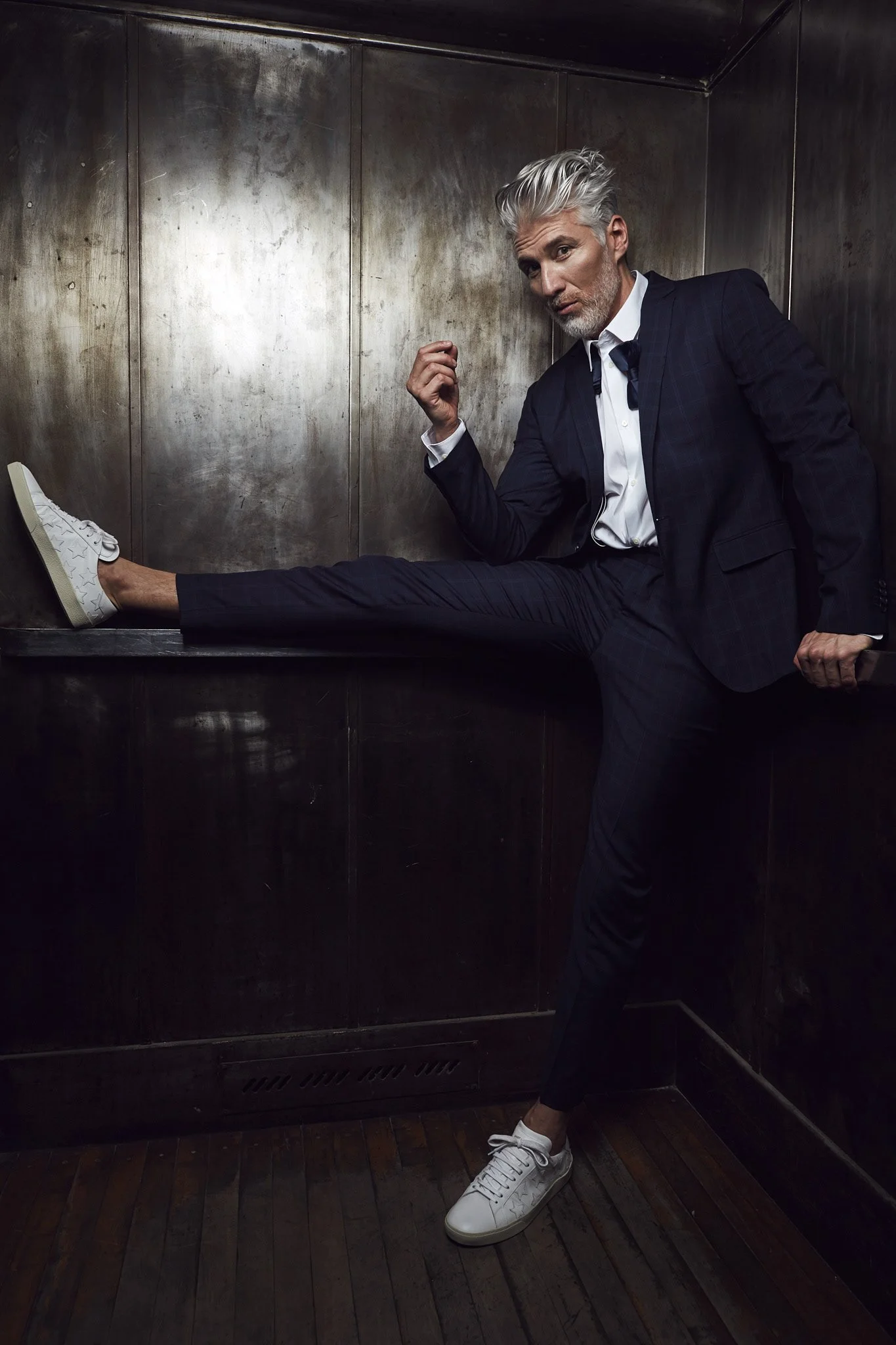 Fashion photography by Justin Munitz - model in a dark suit and white sneakers, seated in a metallic-walled elevator