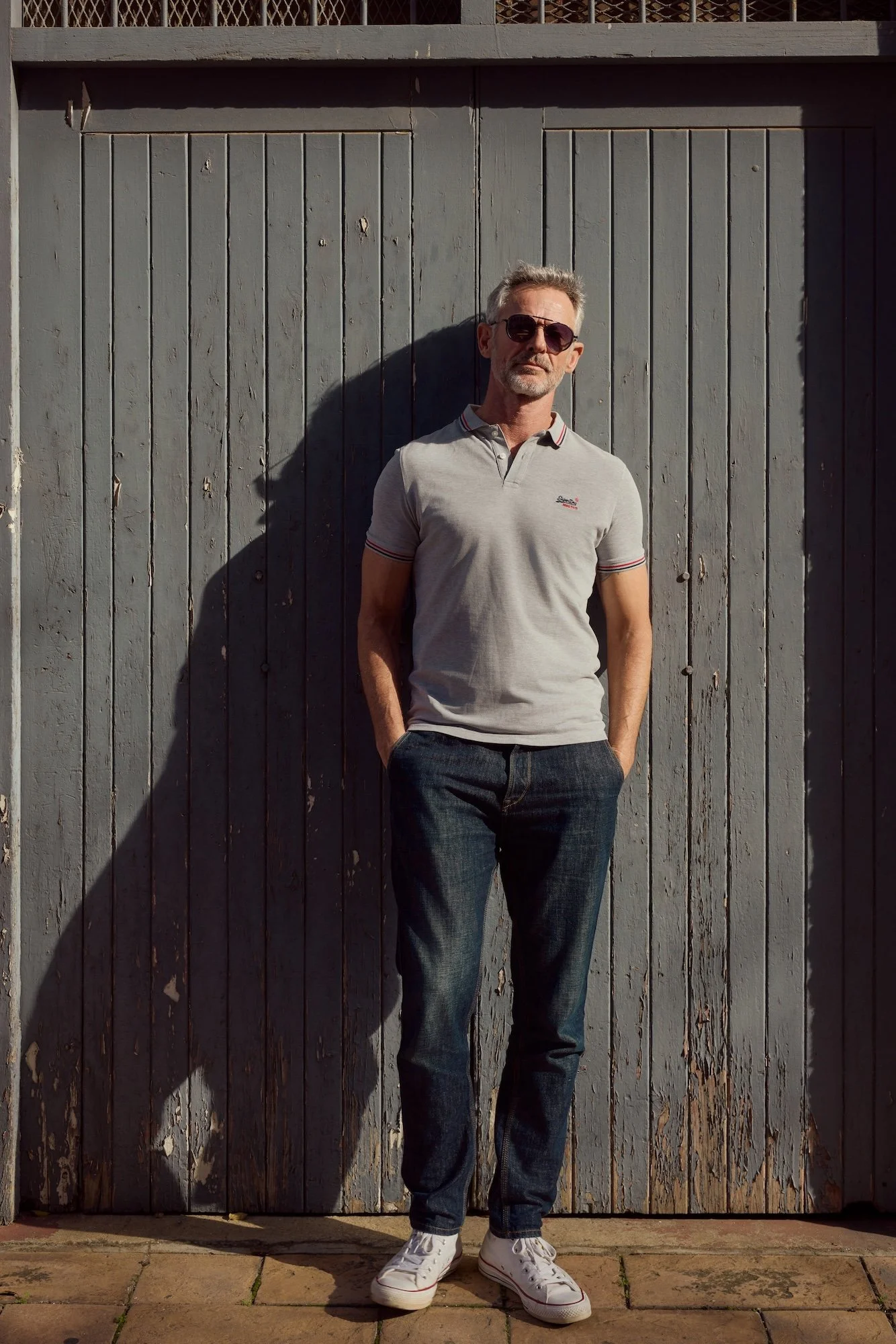 A man wearing sunglasses, a gray polo shirt, blue jeans, and white sneakers, standing in front of a weathered gray wooden fence.