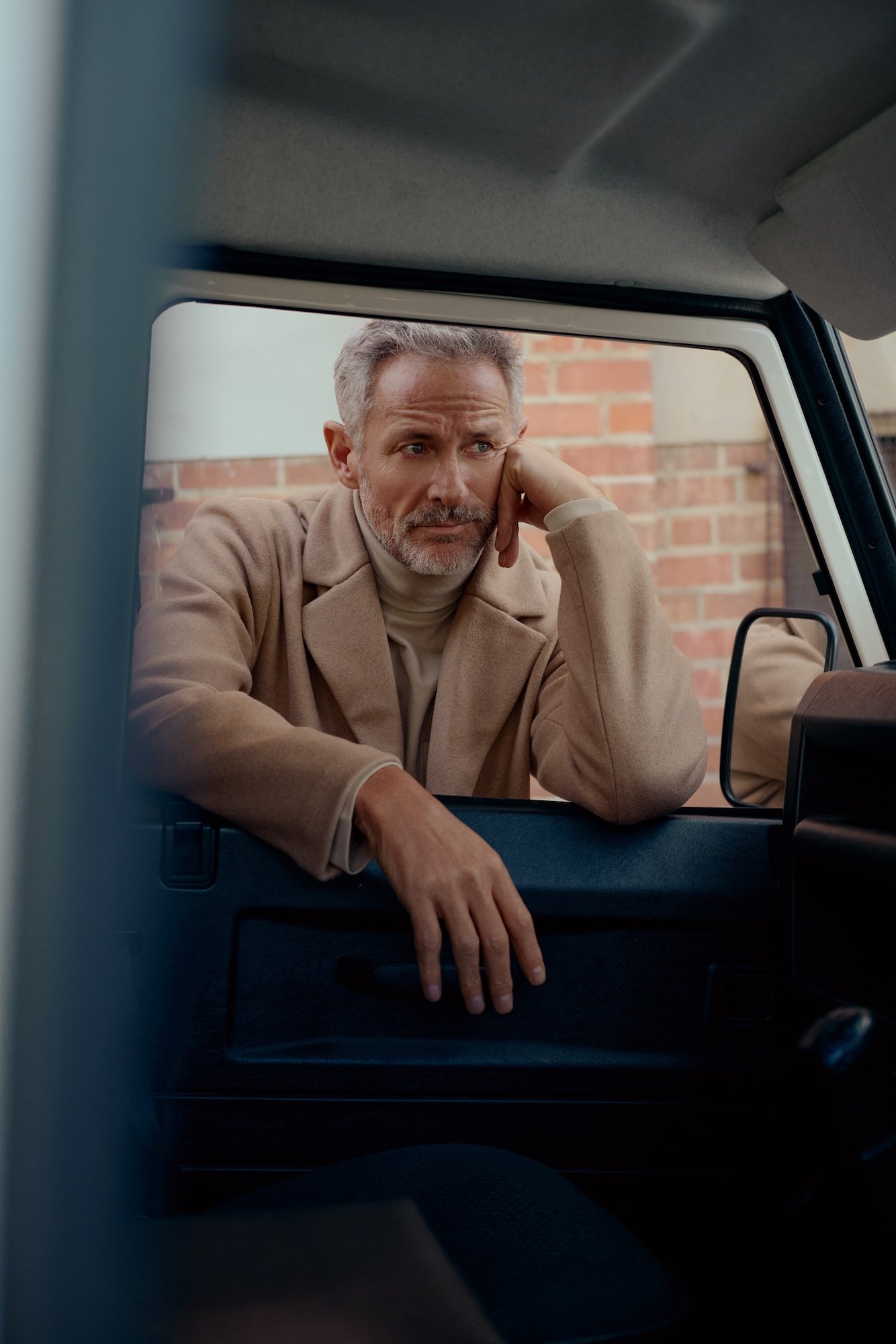 A middle-aged man with gray hair and a beard, wearing a beige coat, leaning on a car window with his hand hanging out, looking pensively.