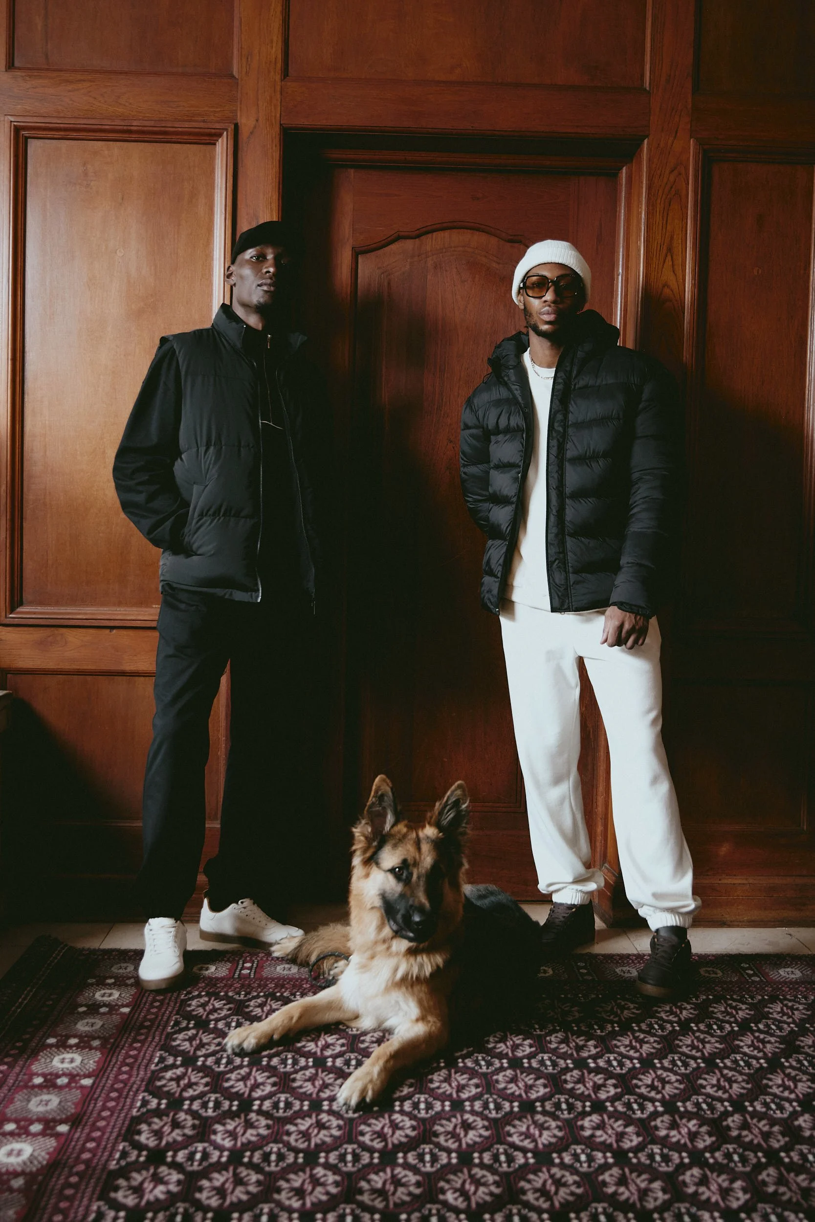  Fashion photography by Justin Munitz — two models in streetwear with a German Shepherd, standing against wood paneling.